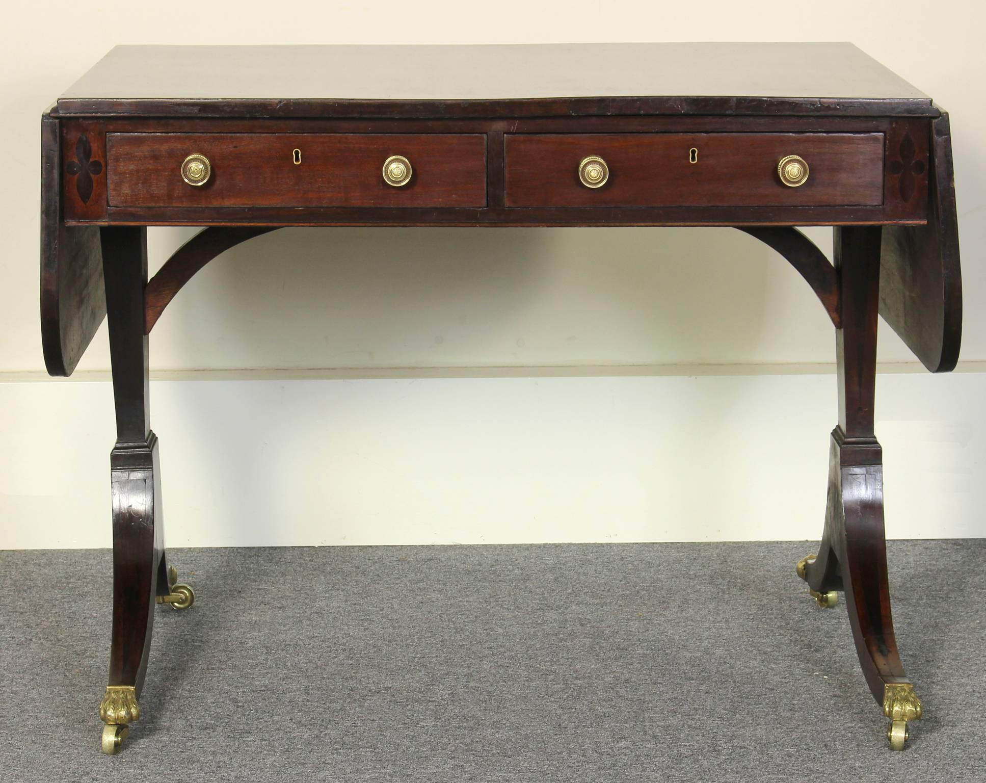 An elegant early 19th C. Regency mahogany sofa table accented with rosewood and ebony trim. The drop-leaf top over two drawers and two dummy drawers, raised on twin end supports and four saber legs terminating in brass lions' paw casters. 