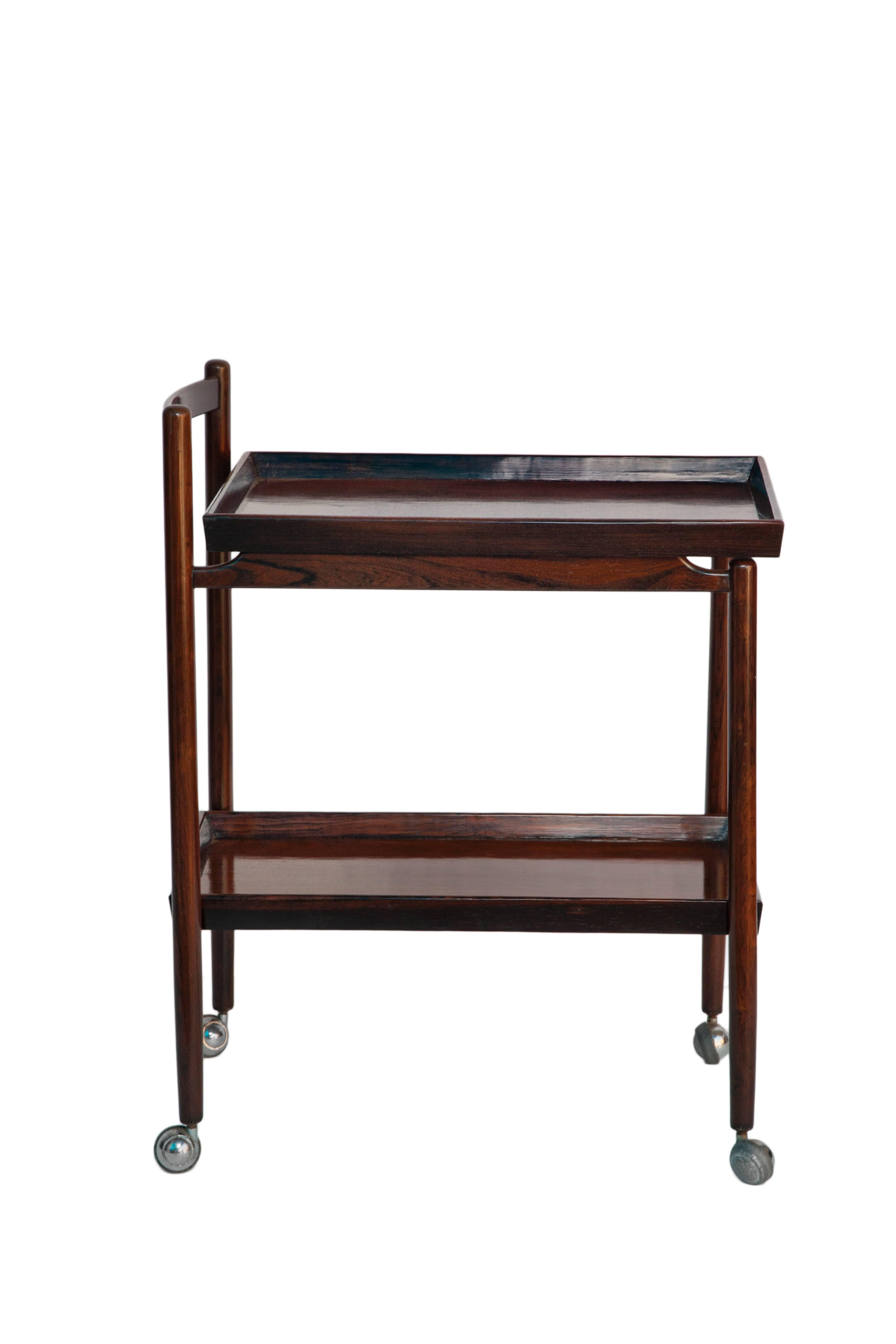 A 1960s two-tier bar cart by designer Sergio Rodrigues, entirely in rich Brazilian wood, on metal caster wheels. Top tray is removable. Excellent vintage condition, with minuscule patina to metal, consistent with age.
