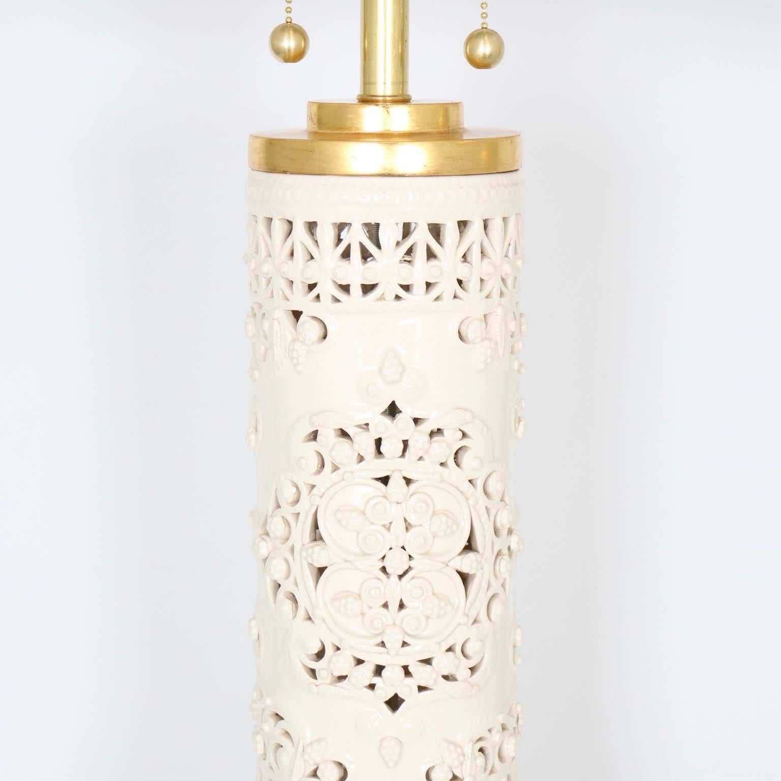 A pair of Hollywood Regency style lamps, produced in Italy, circa 1960s in white ceramic with intricate pierced pattern, caps and bases in gilded wood. The noted height is to the finial. The height to the top of each ceramic body is 25.5″. Fully