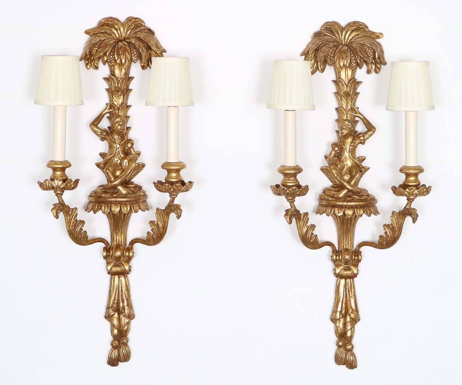 A pair of Italian Mid-Century Modern era 1950s candelabra wall sconces, influenced by the Hollywood Regency style, each of ornately carved gilded wood, featuring monkeys seated beneath palm trees, flanked by two-lights with acanthus arms. Wiring and