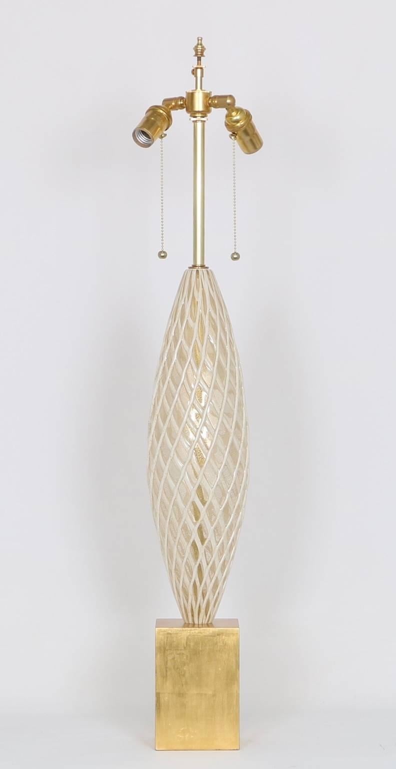 This circa 1950s lamp by Venini is a magnificent sample of Italian Murano art glass, with reticulated white and gold ribbons over clear glass body with gold flakes, mounted on a gilded wood base. The noted height is to the finial. The height to the