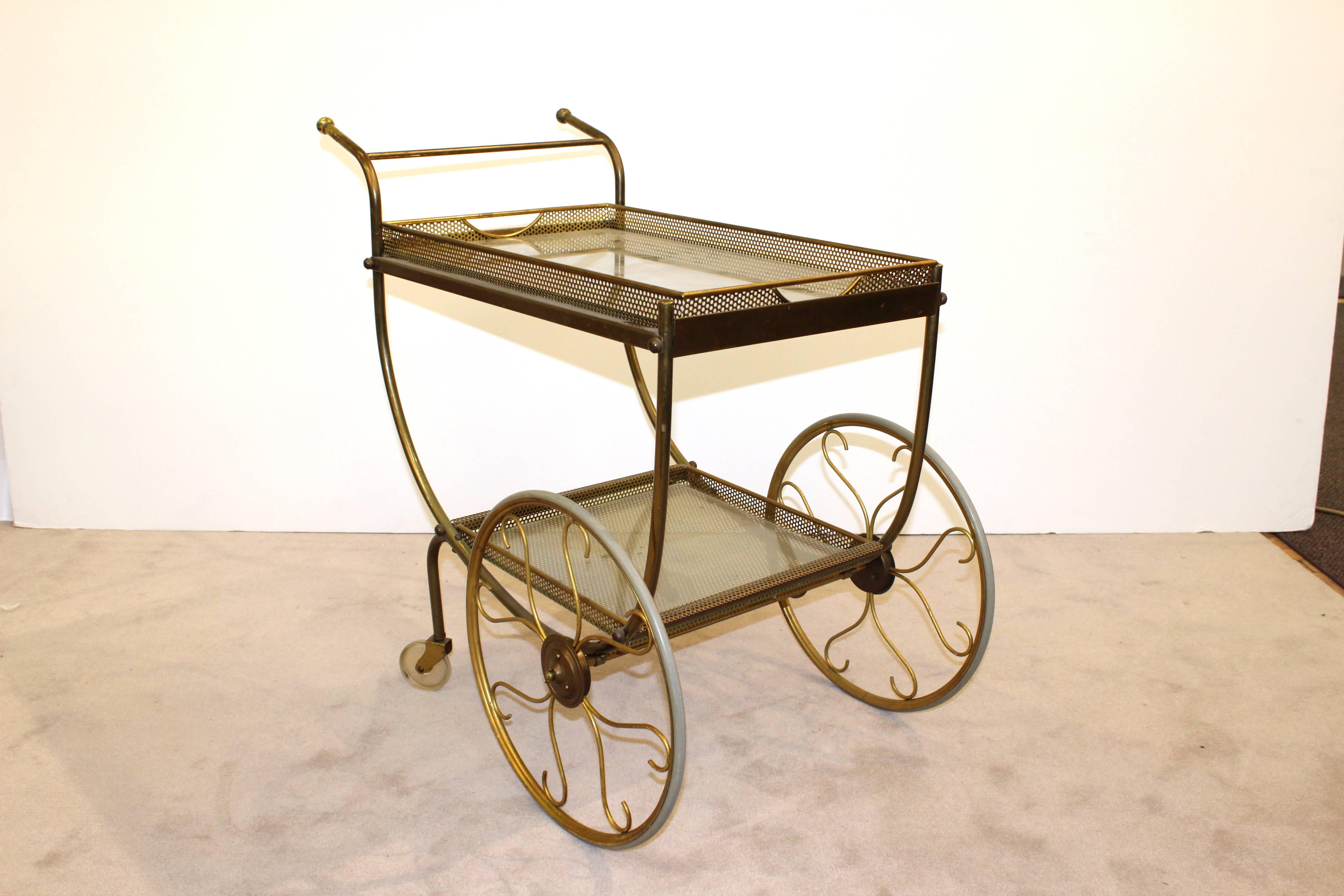 This bar cart, dating from the mid-20th century features two perforated metal tiers, each lined in glass and four wheels. The spokes on the set of the two larger wheels are curved. The top tier of the bar cart features a removable tray with two