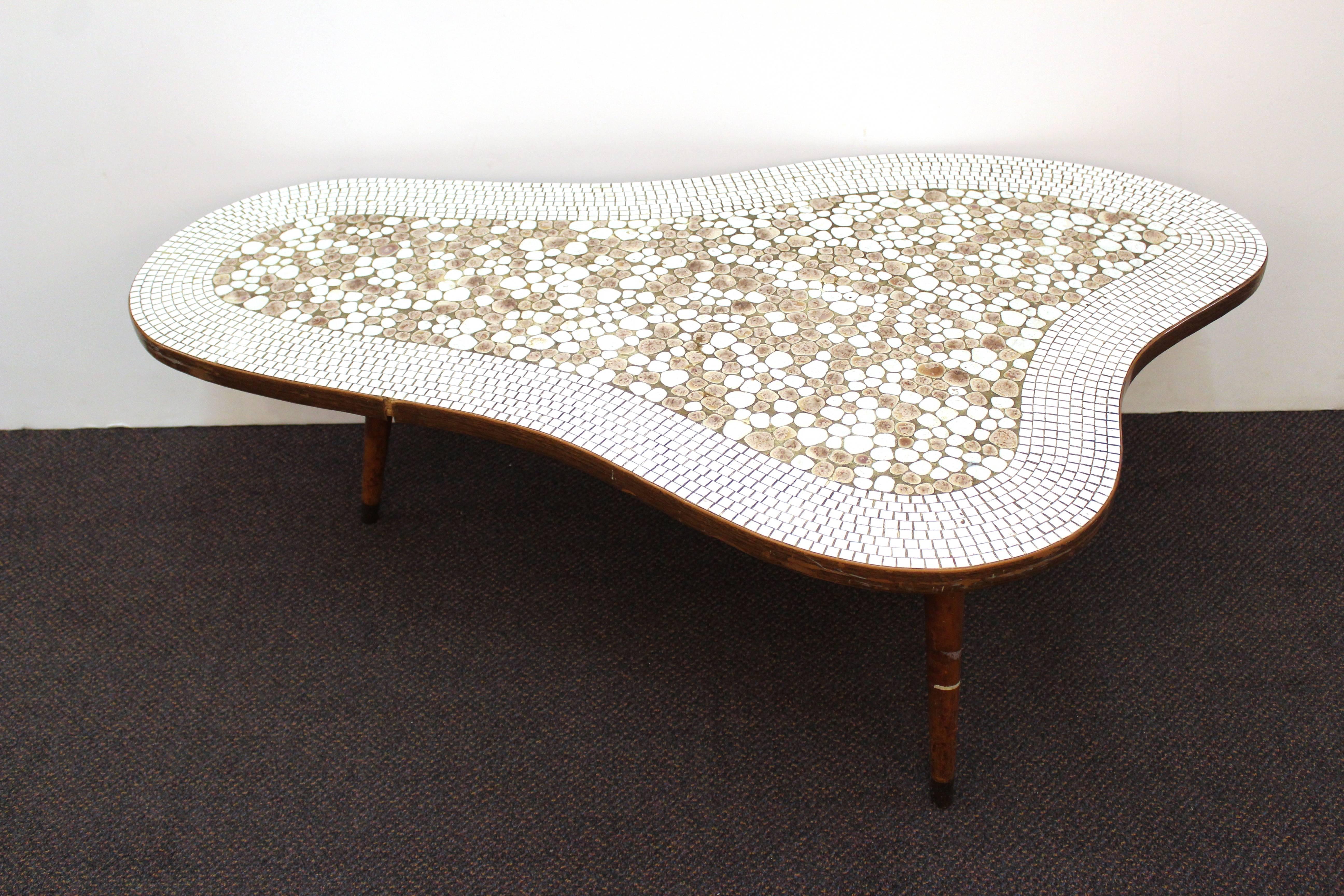 This Mid-Century coffee table features a mosaic stone top in white and grey. It is in an organic form and rests on three wooden legs. There are a few cracks and scuffs but this table is in overall good vintage condition. 110908.