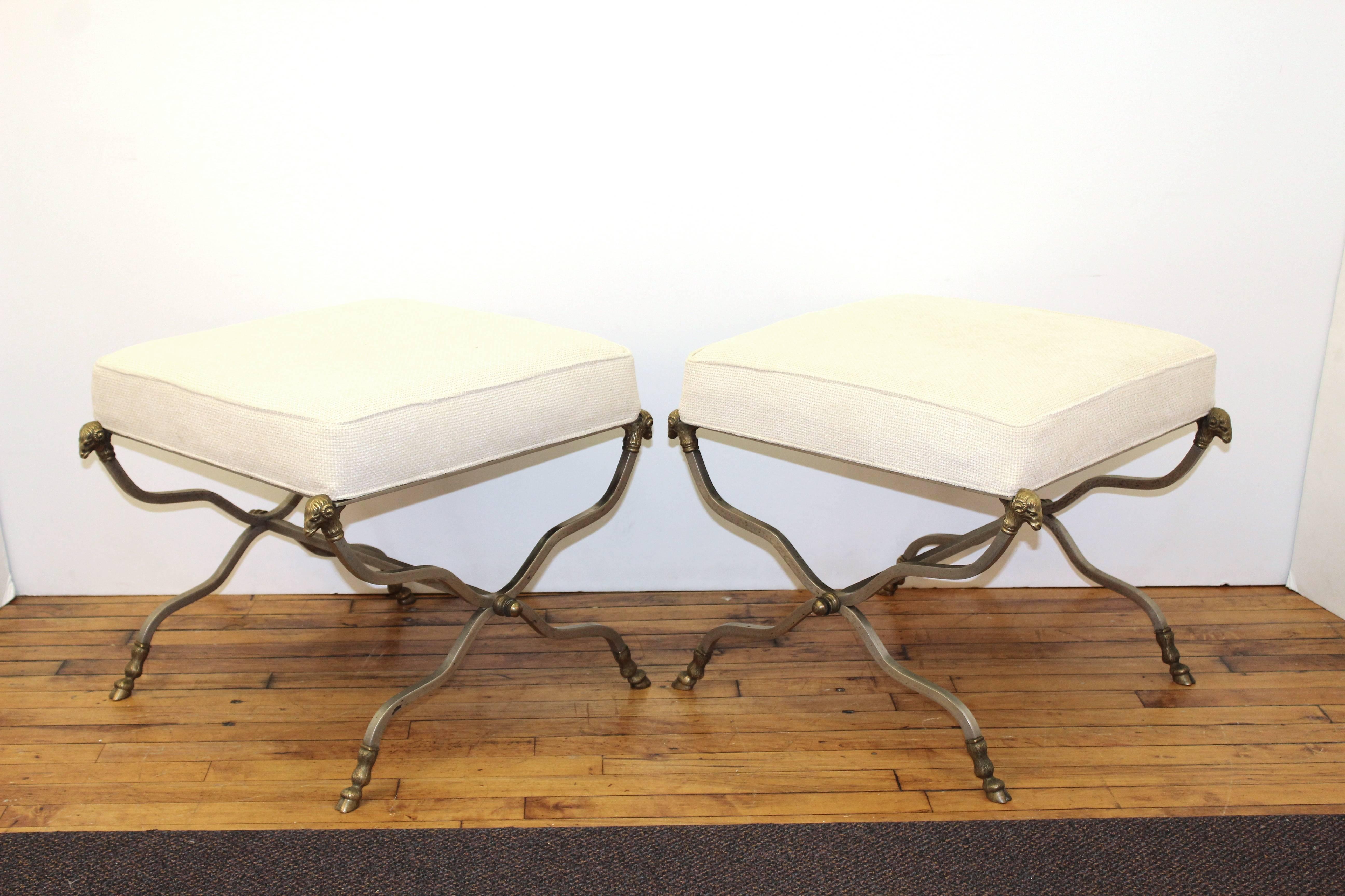 Hollywood Regency Maison Jansen Attributed Curule benches in metal with ram's heads adorning the upper part of the frame and ram's hooves adorning the feet. Each bench features a light cream upholstery and is in good vintage condition, consistent