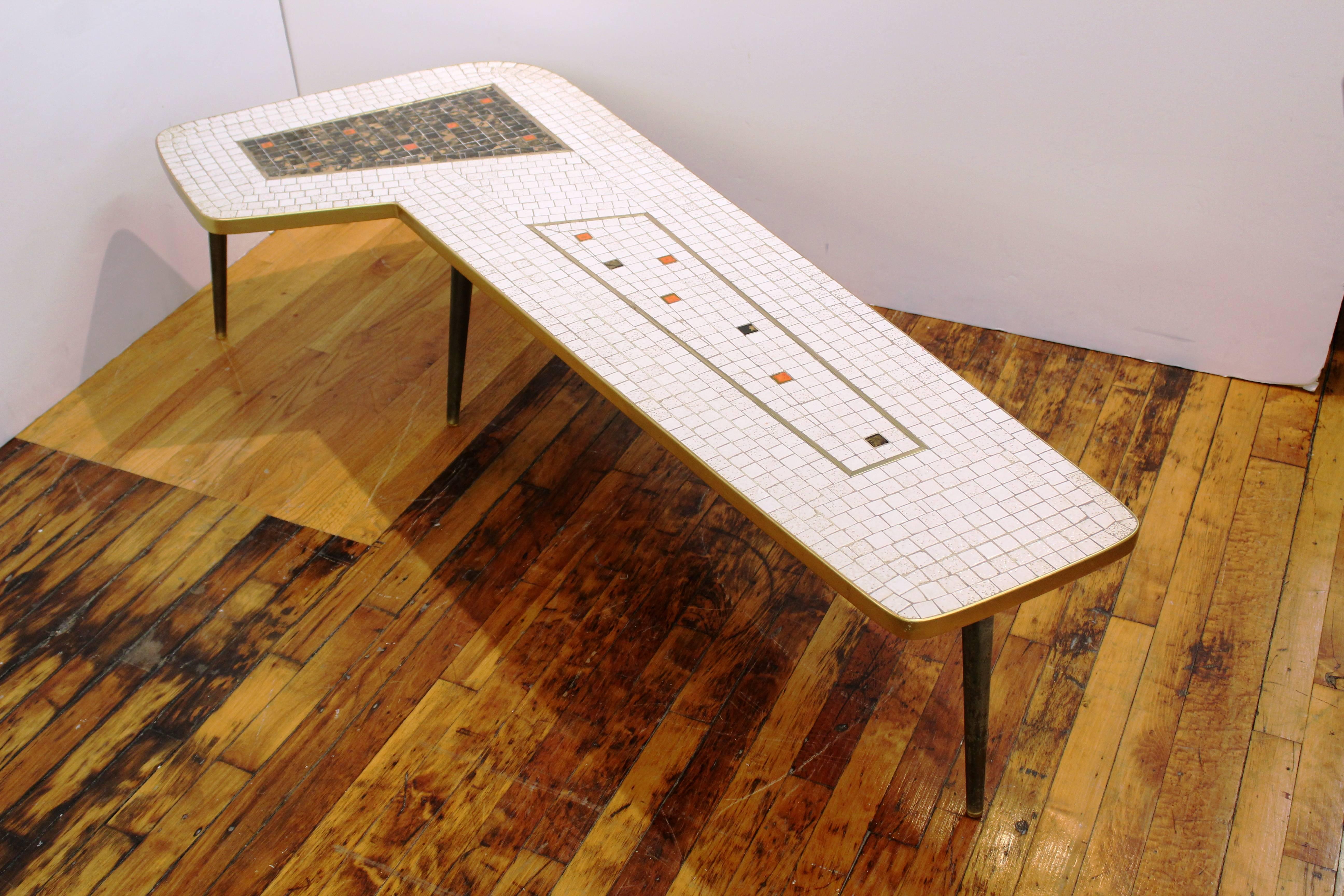 A Googie style L-shaped coffee table with mosaic space-age top and brass tapered legs. The top features white, red, gold tone and black tiles while the wooden legs are painted black. In very good vintage condition having wear consistent with age and