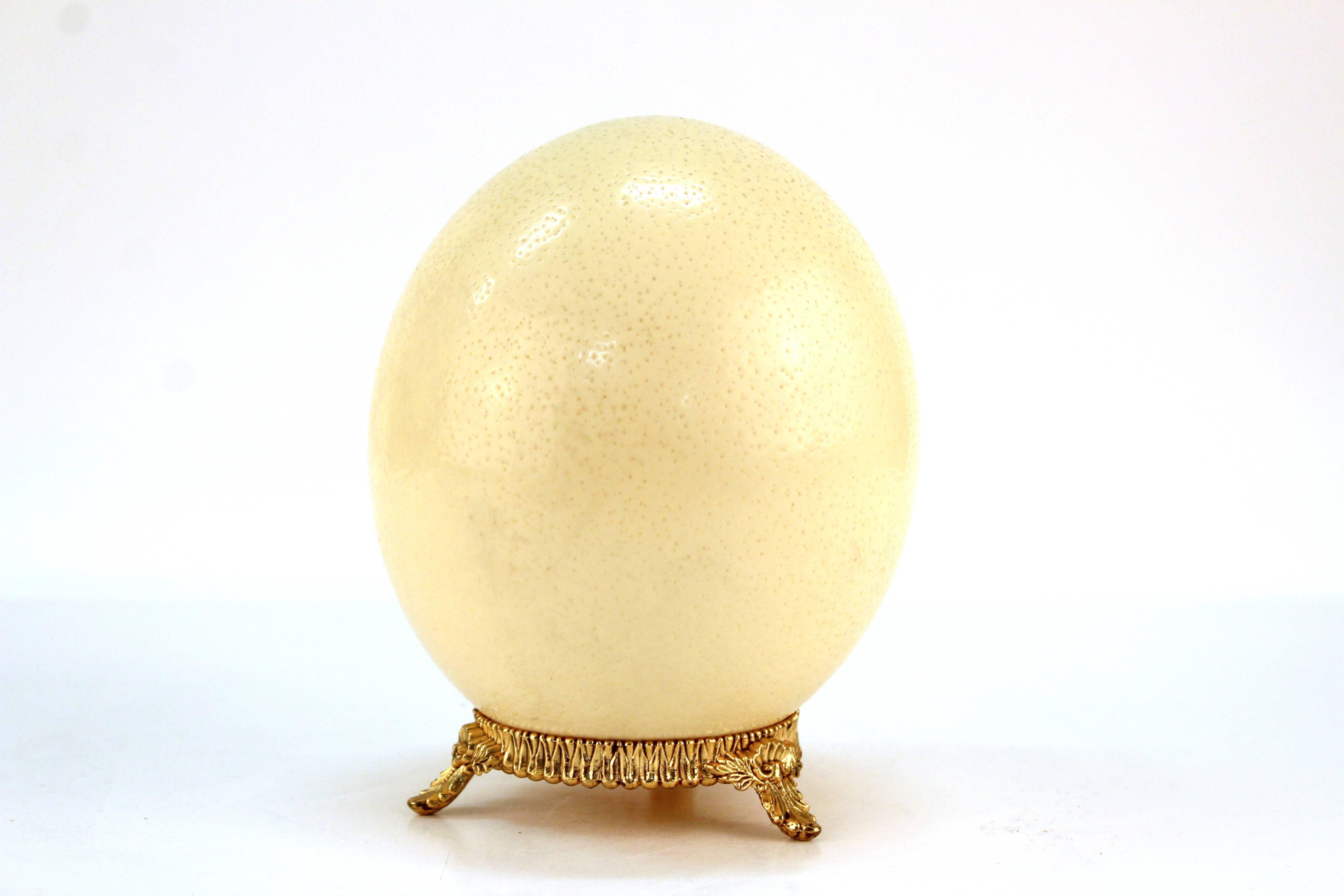 An antique ostrich egg placed atop a heavily embellished stand. The gold-tone metal egg mount stands on three floral inspired legs. In very good vintage condition.
