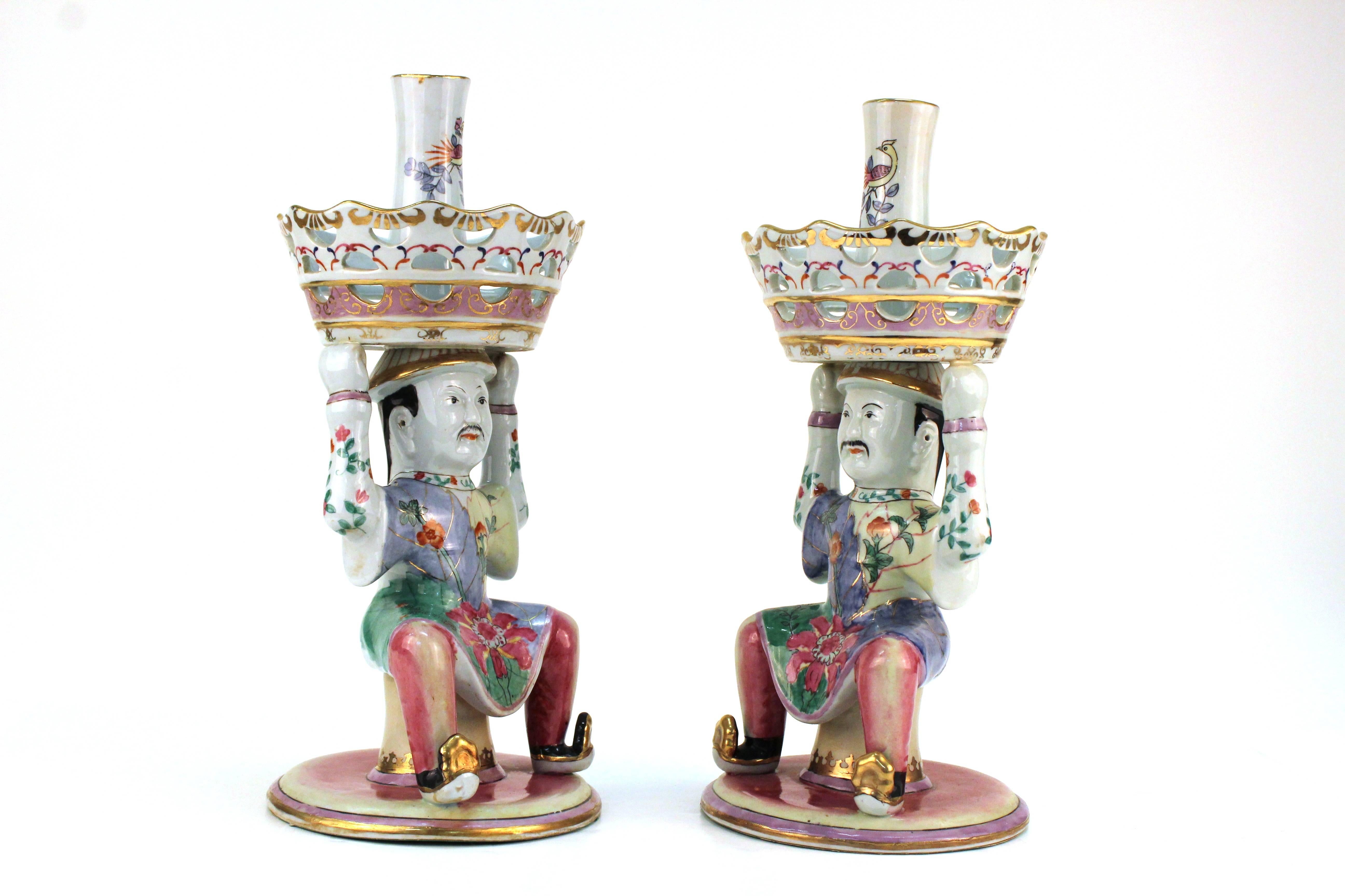 A pair of epergnes with baskets held by Chinese men in brightly colored tobacco leaf pattern robes. The tops include geometric cut-out and are painted with florals patterns with gold-tone accents. The bottom of both ceramic figures are covered in