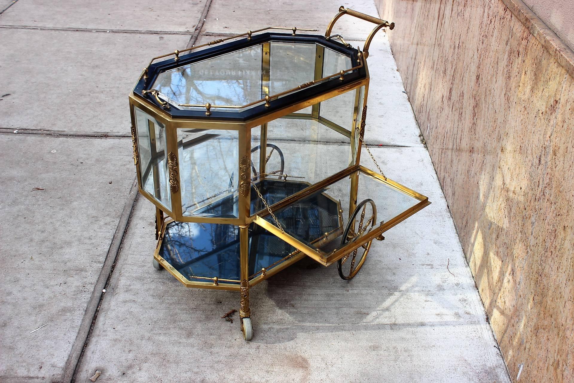 A brass tea trolley or bar cart with upper and lower galleries, glass-topped trays and accented ormolu mounts. The top with ebonized wood trim and brass gallery is a large removable tray. The lower galleried tray is fixed in place and features clear