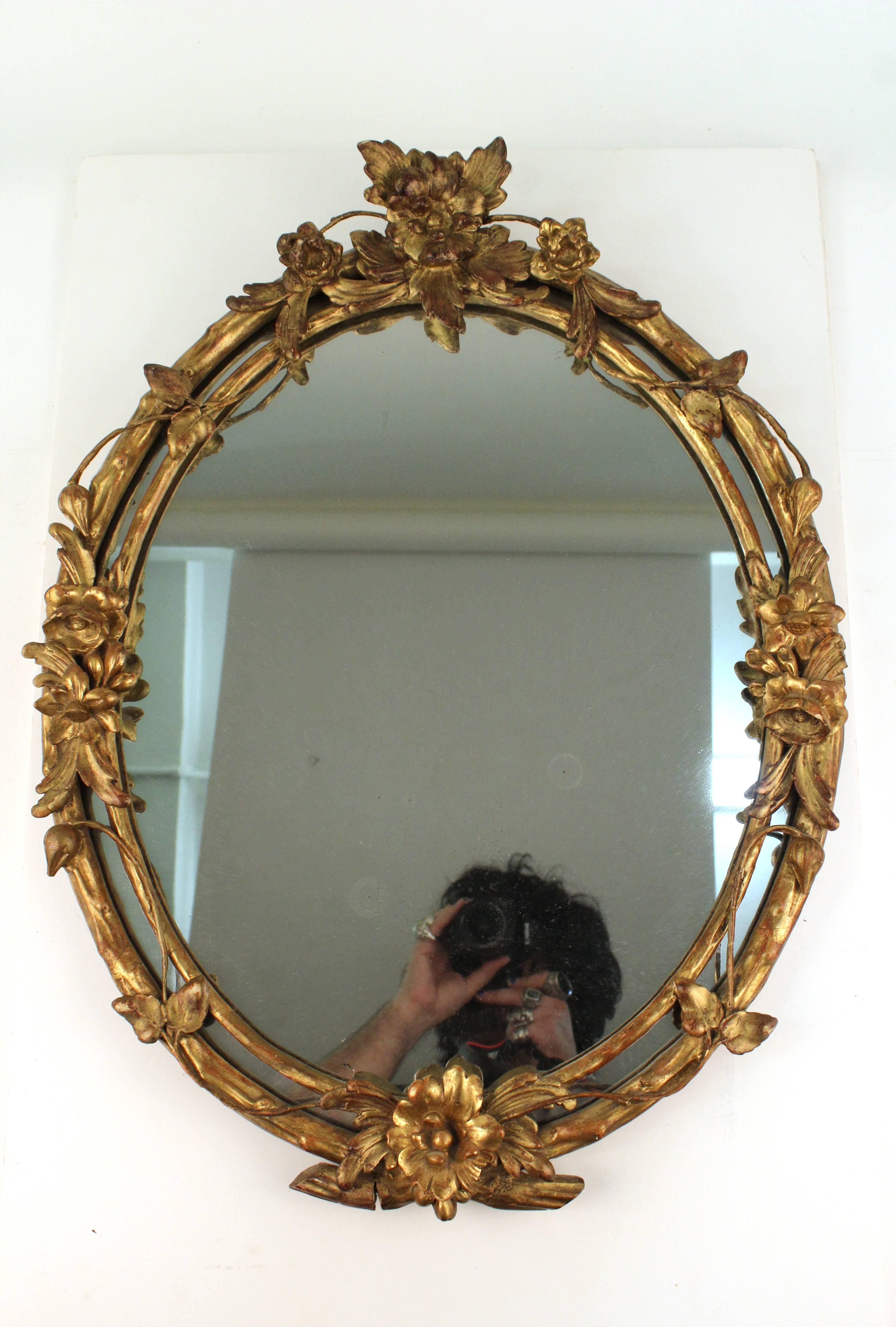 A giltwood mirror in a carved oval frame. Crafted to look like a double frame connected by winding vine with flowers. Includes the original label stating [Made in France]. Some wear appropriate to age and use, namely some chips in the gilt and wear