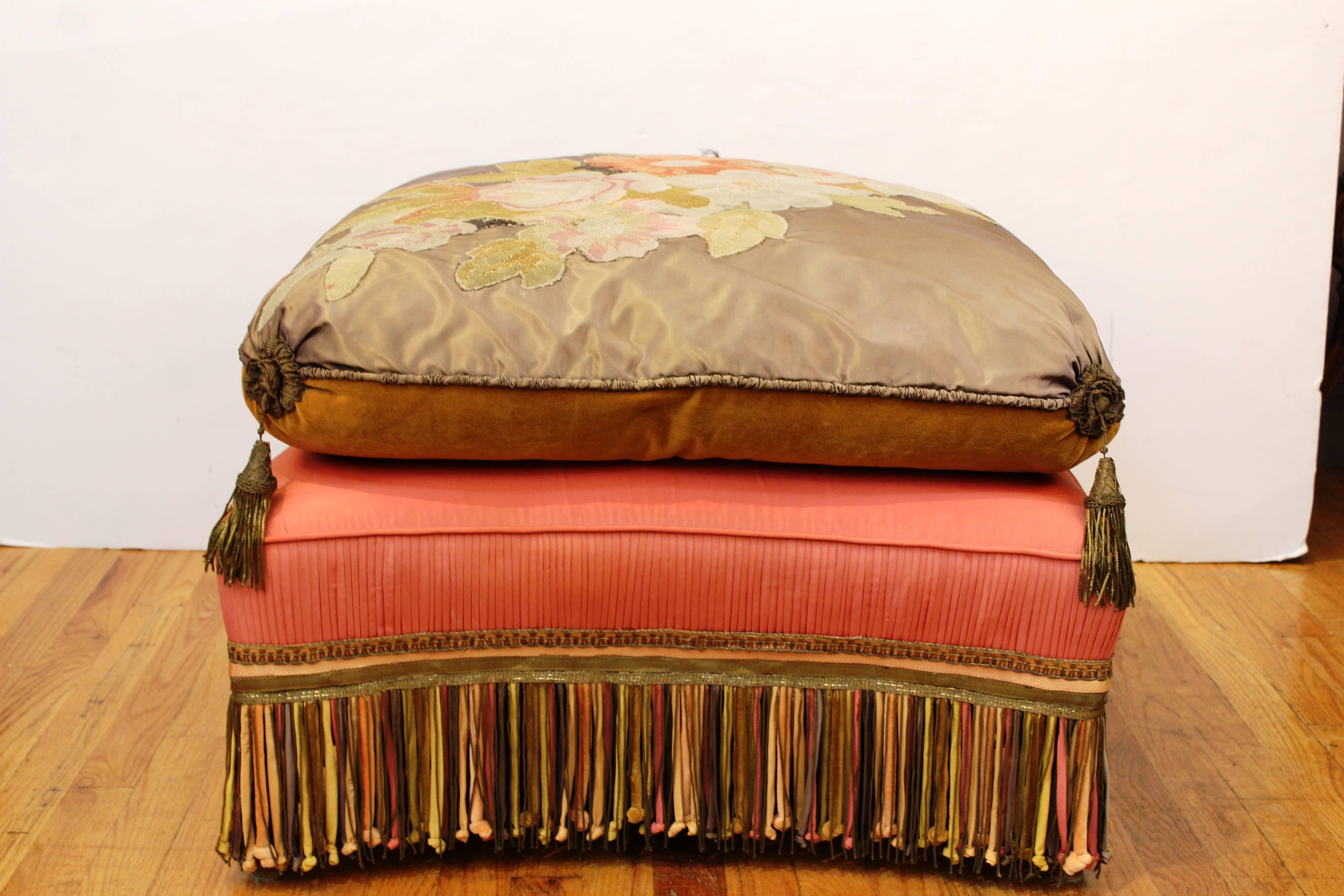 A large Victorian ottoman with detailed floral needlework on the seat cushion. decorative borders and tassels at the corners, with a refined multicolored fringe. Wear appropriate to age and use, in good condition.