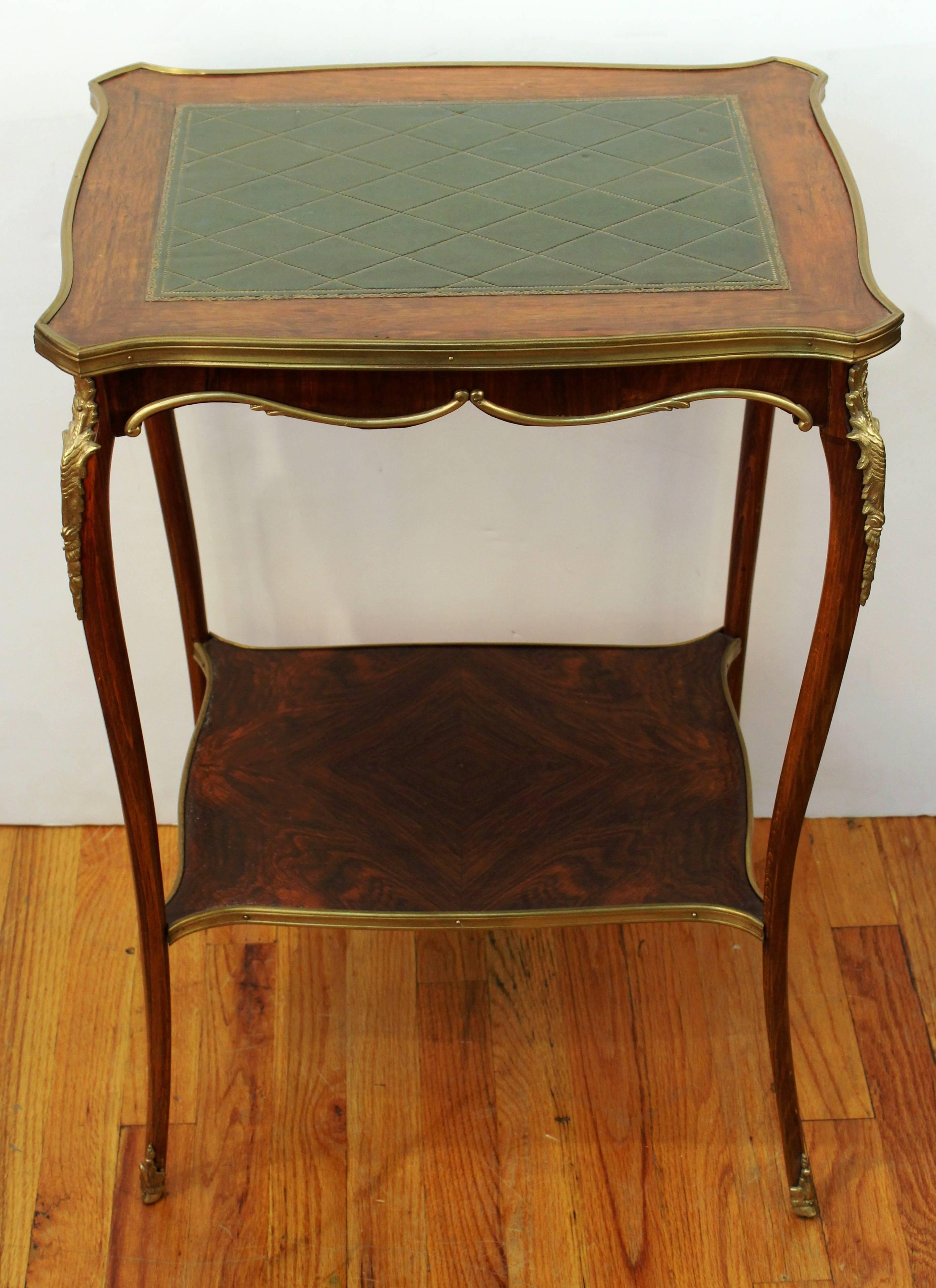 Wooden table dating from the 19th century with a lower shelf. The tabletop features green leather and the sides and corners are accented with decorative brass pieces. This table is in good vintage condition consistent with age and use. There are a