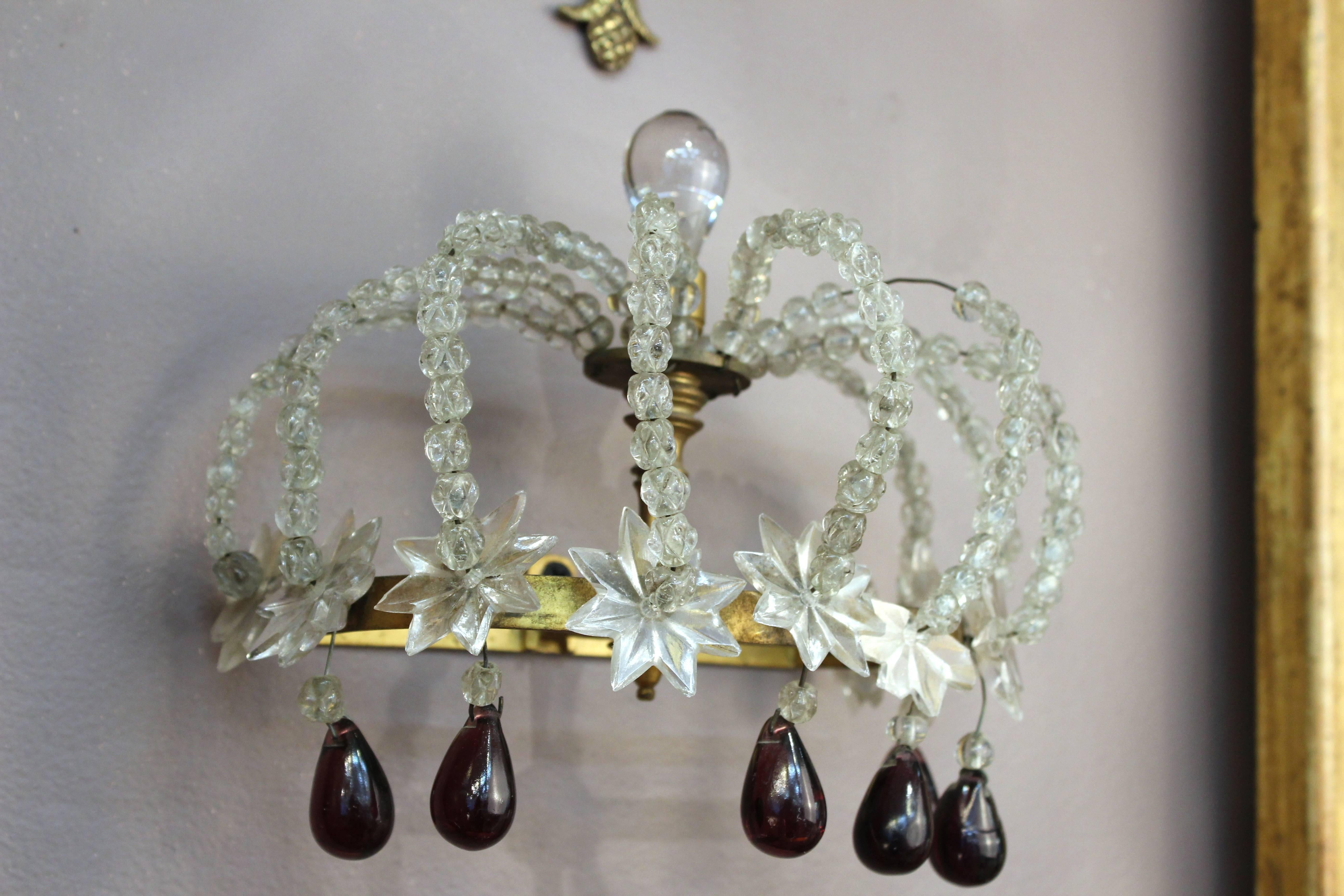 A pair of sconces in the shape of crowns. Crafted in clear rock crystal beads on brass base with stars on the band and dangling plum colored drops. Purely decorative, the pieces do not hold candles or include lights. Despite a missing drop on one