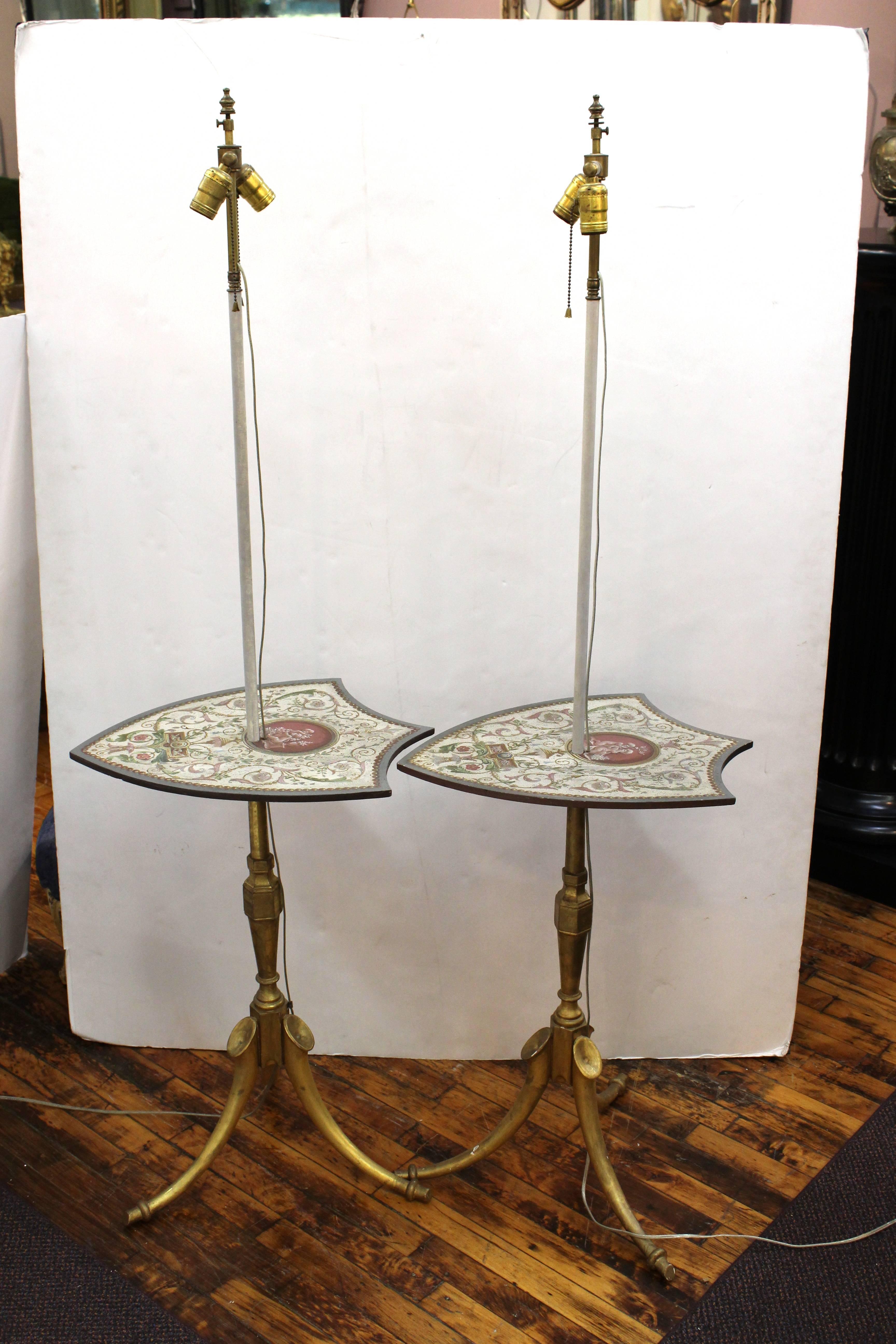 A pair of floor lamps crafted from antique shield shaped firescreens; possibly of Russian origin. The shields are painted with scrolls, flowers and putti. Each lamp stands on a giltwood tripod base made up of three Horn forms. Some wear appropriate