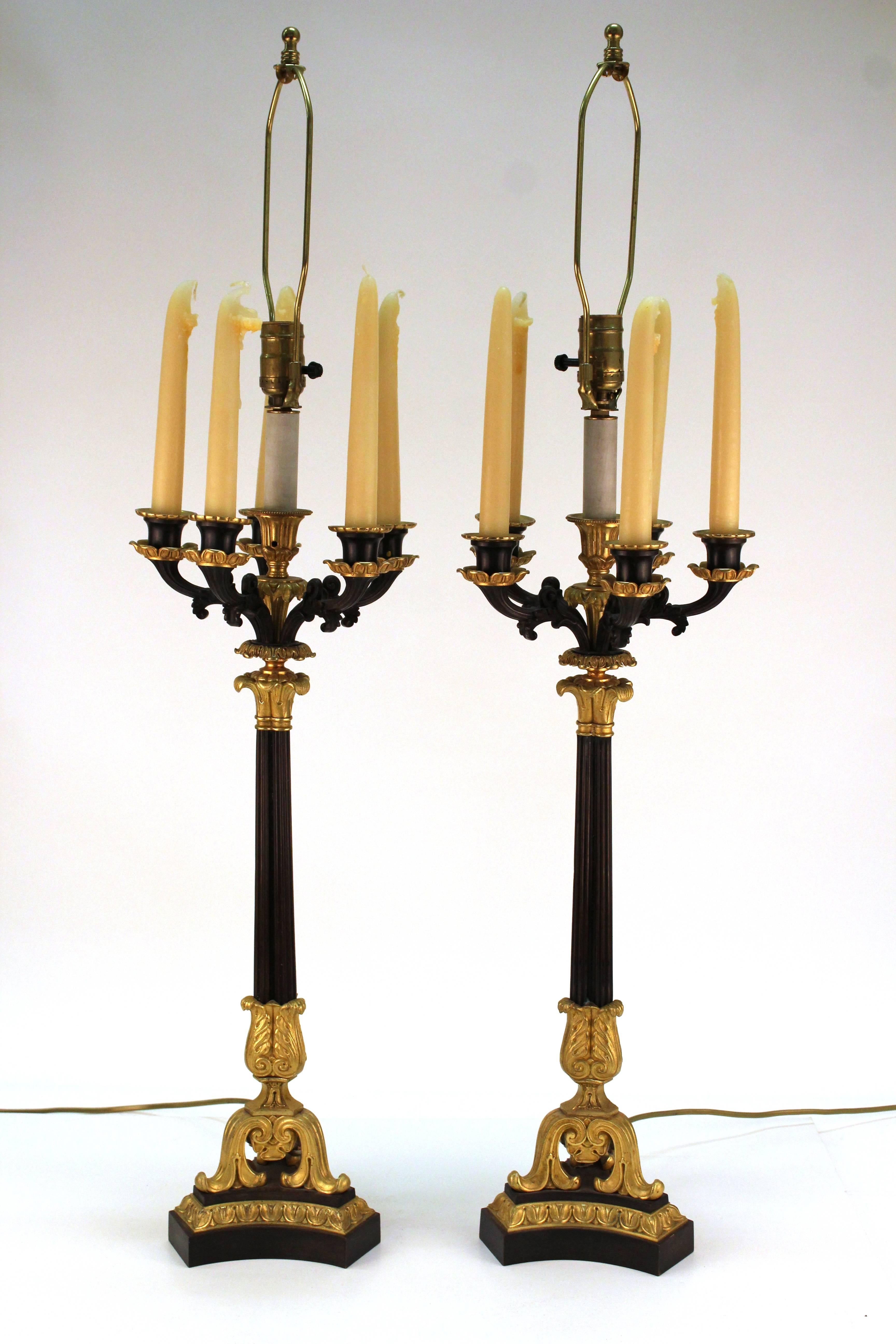 A pair of Empire style table lamps in the form of candelabras produced in France. Both pieces include five candle holders at the end of scrolled arms; shown here with real candles. The lamps are bronzed with gilt acanthus leaf accents. Both are in