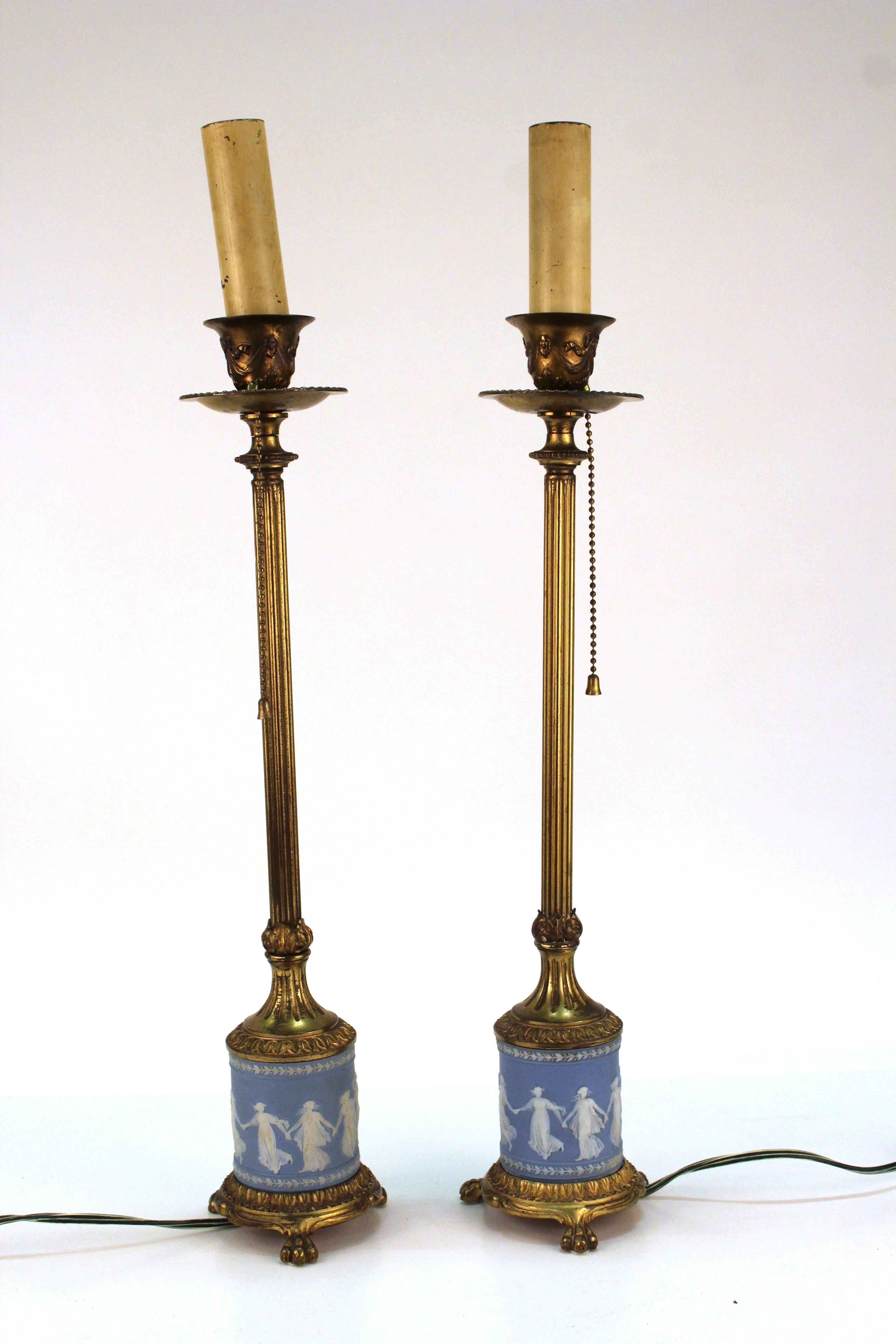 A pair of table lamps with wedgwood base. The lamps are gilt and stand on three clawed feet. Decorative details include long skinny column necks, acanthus leaves and bows along the top. The lamps are topped with faux candle sockets. Despite minor