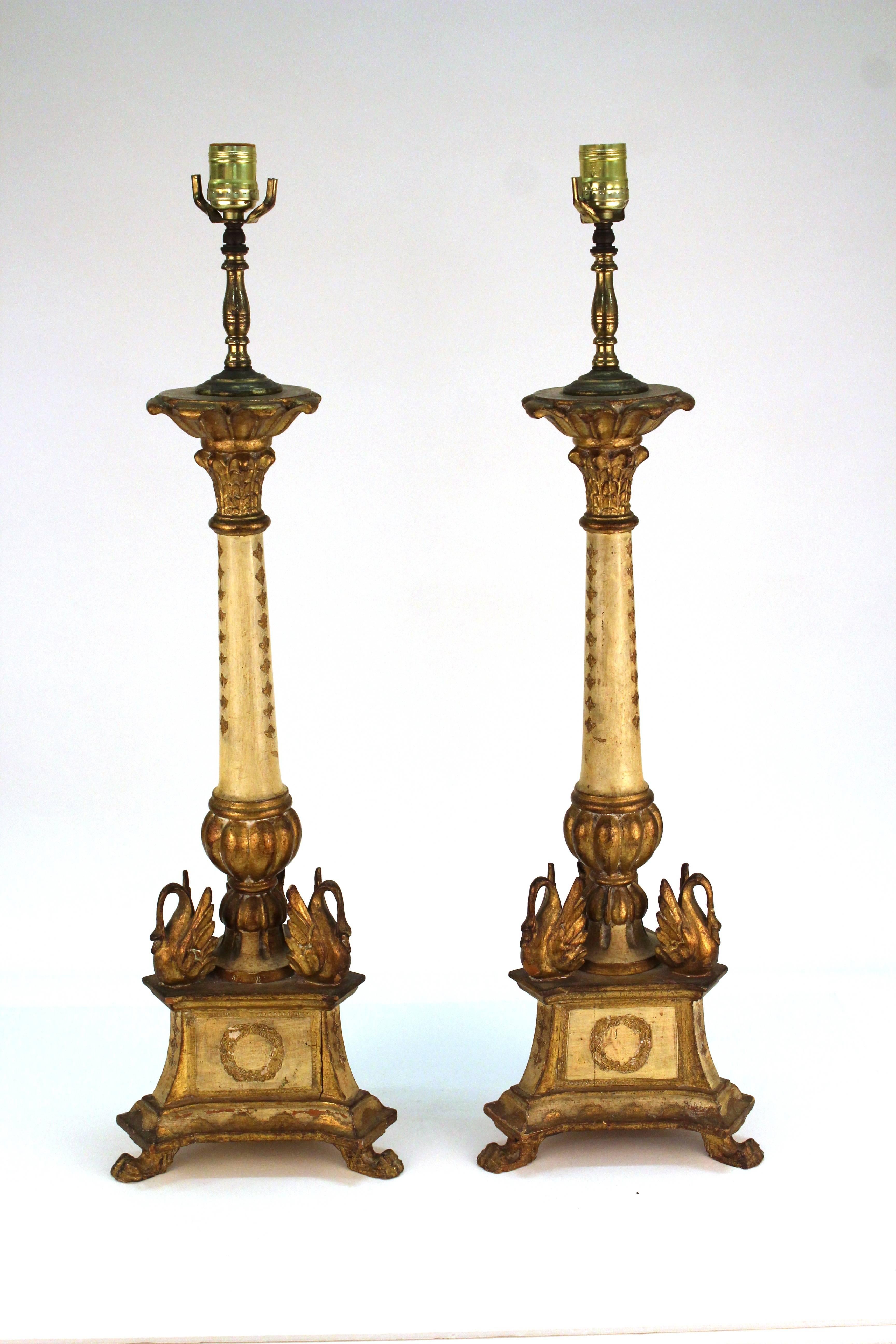 A pair of torchere table lamps with swans along the base. The lamps stand on three clawed feet that support a trunk decorated in wreaths. Marked [M. Italy] on the bottom. The pair are giltwood with minor wear appropriate to age and use. The lamps