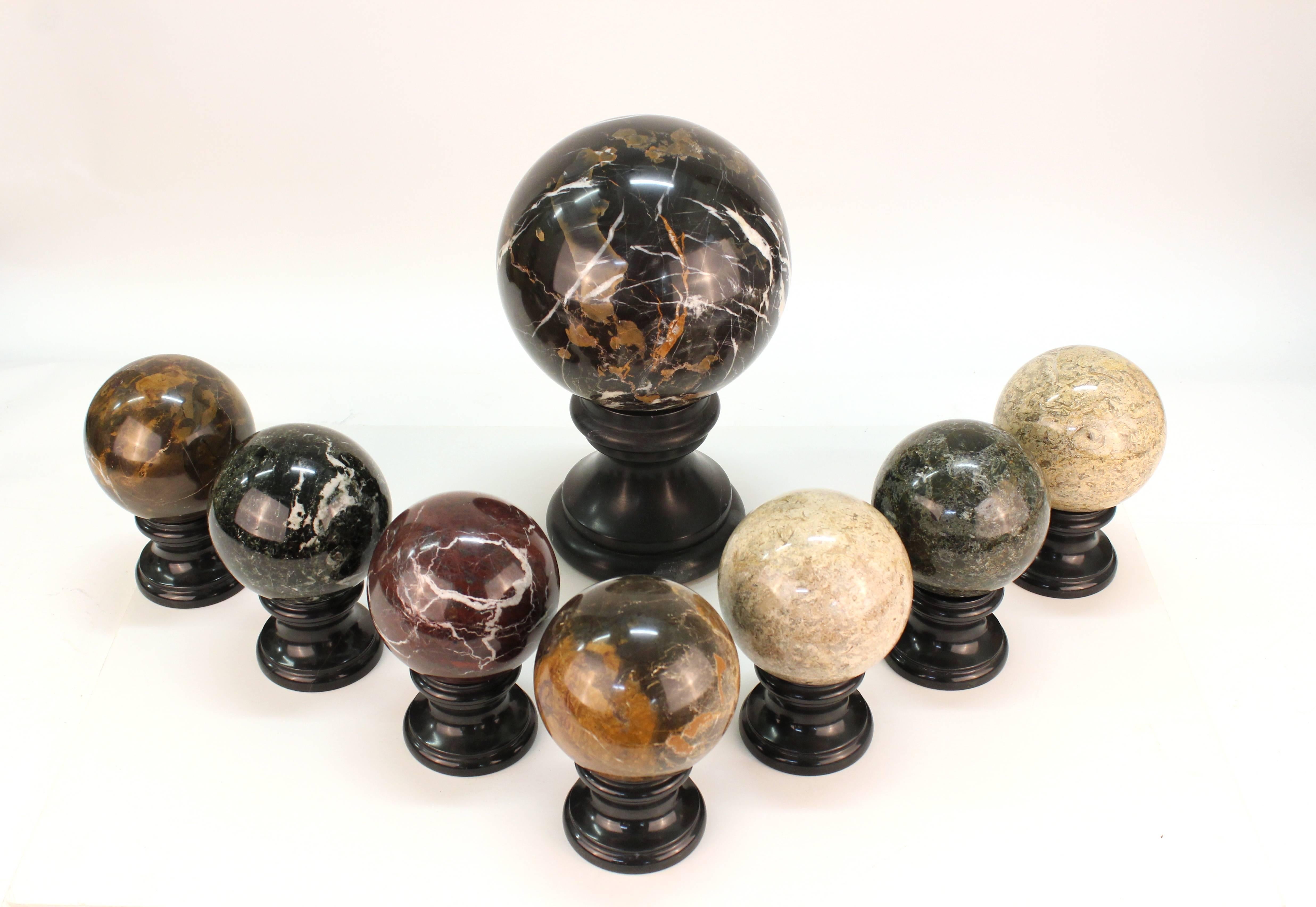 A set of of decorative marble and stone balls. All in various shades of green, red, beige, black and yellow. The set includes one large ball and seven smaller ones all mounted on black stone bases. Each small ball has a similar colored pair except