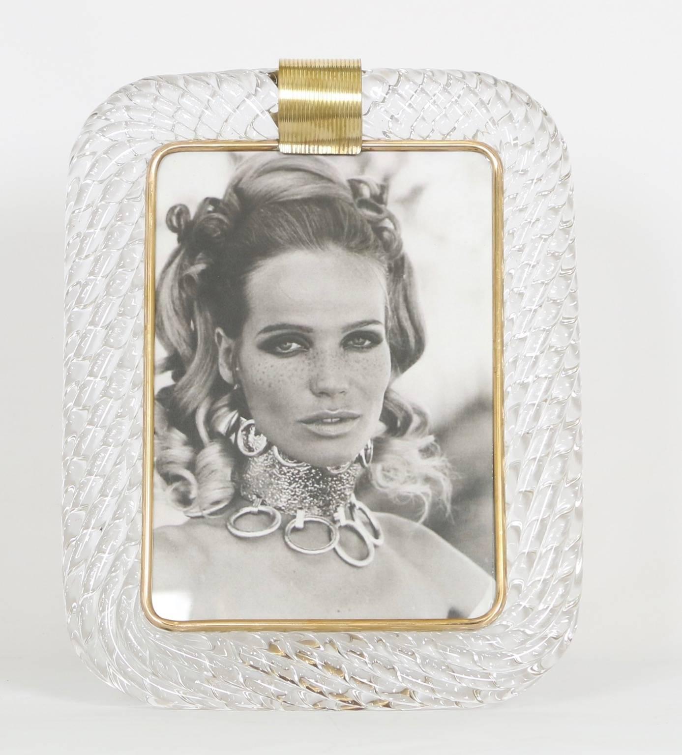 A large Venini photo frame in clear Murano glass. The top of the frame features a band of ribbed brass as an accent as well as a brass band around the inner edge of the frame. There is a brass stand attached to the back. This piece is in excellent
