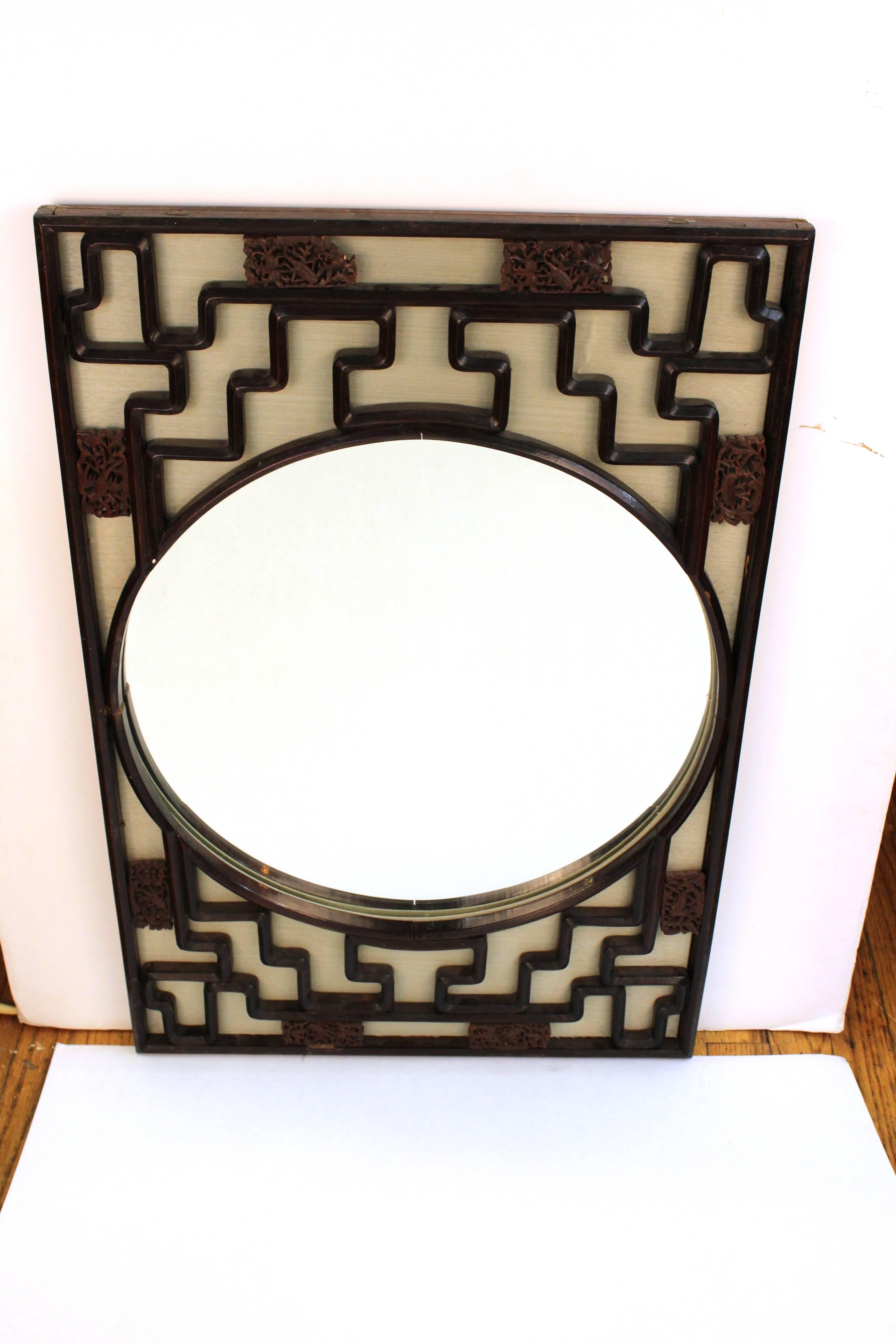 A round wall mirror from Gump's in an Asian inspired Chinese Chippendale style frame. The dark wood frame features cutouts that make geometric patterns as well as intricately carved decorative segments against a light grey woven textile mounted