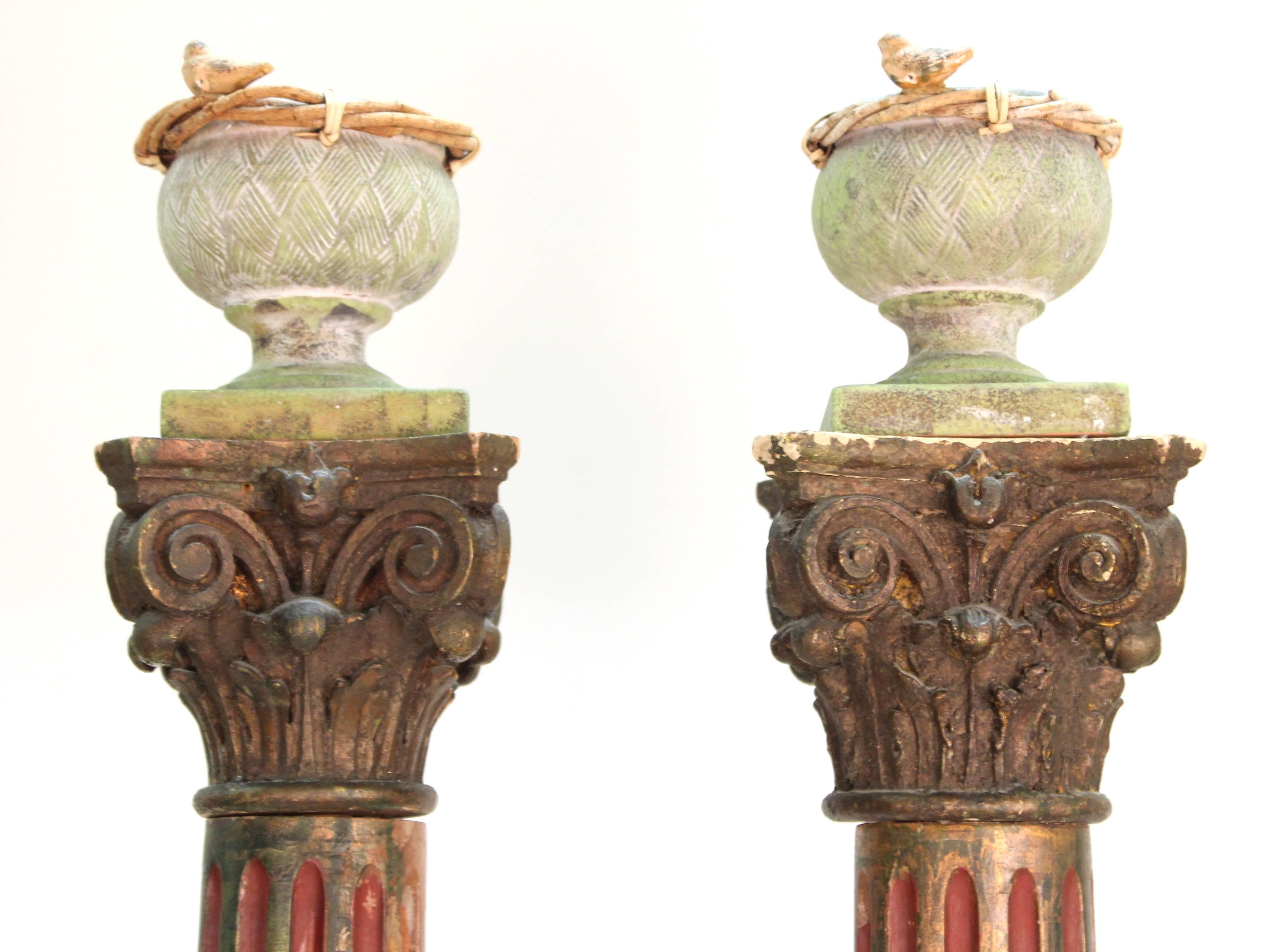 Painted wooden Victorian columns in the Corinthian style with bird nests on top feature black square bases, a red and black fluted shaft, and black capital with curling acanthus leaves. Each column is topped with a ceramic green urn with bird