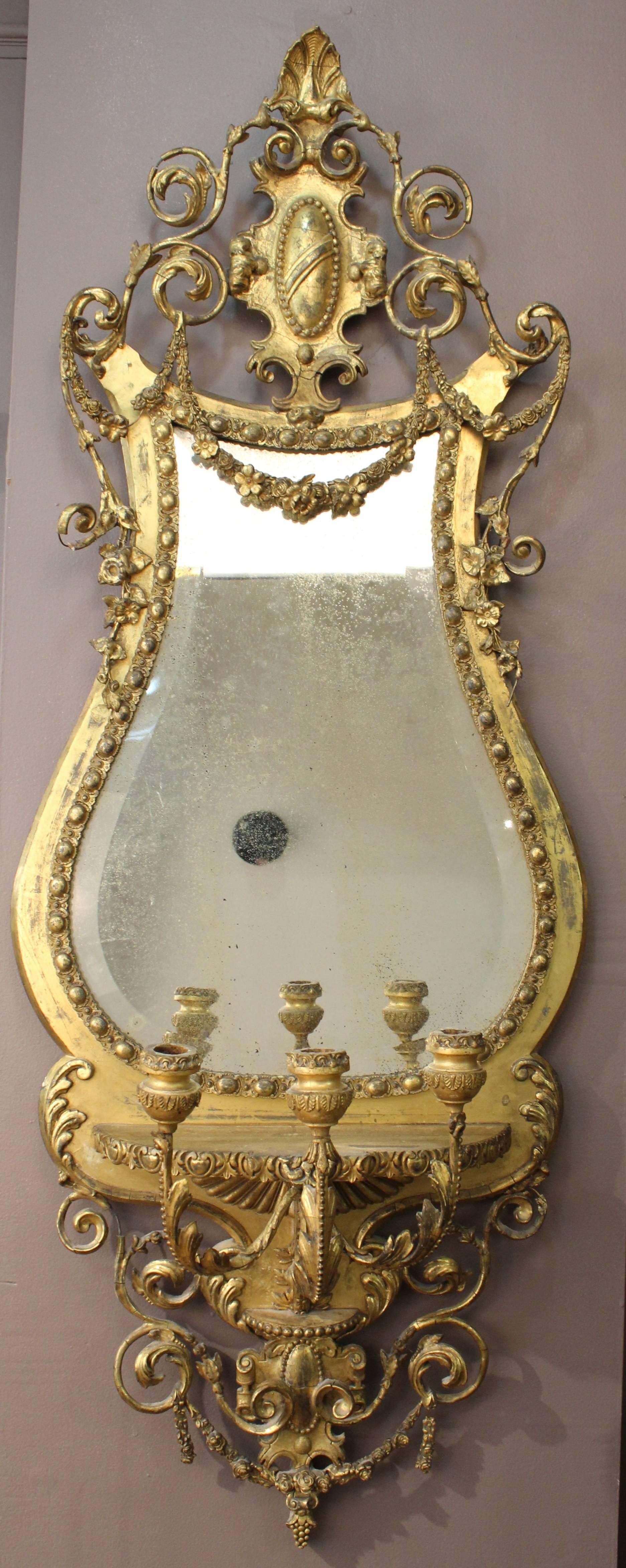 George III period wall mirror in gesso and giltwood. The mirror features ornately carved foliage and a lyre-shaped frame with gilt swags encasing an antique mirror with original patina. The base section contains three candelabra as well as more