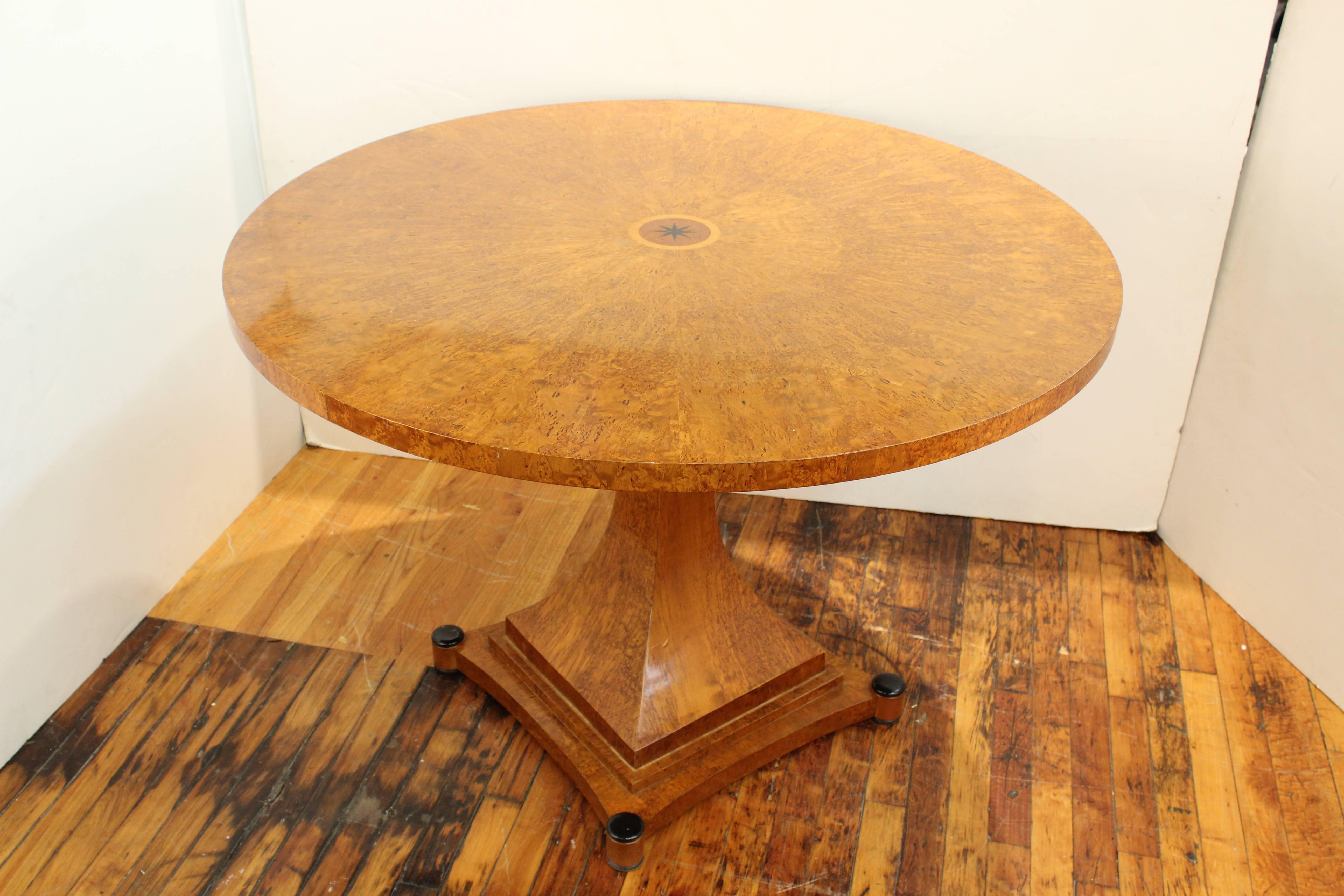 A dining table from the Art Deco period. Crafted in burl wood with geometric detail. Features a round face on a single leg atop a stepped square base. Wear consistent with age and use. The table is in good condition.