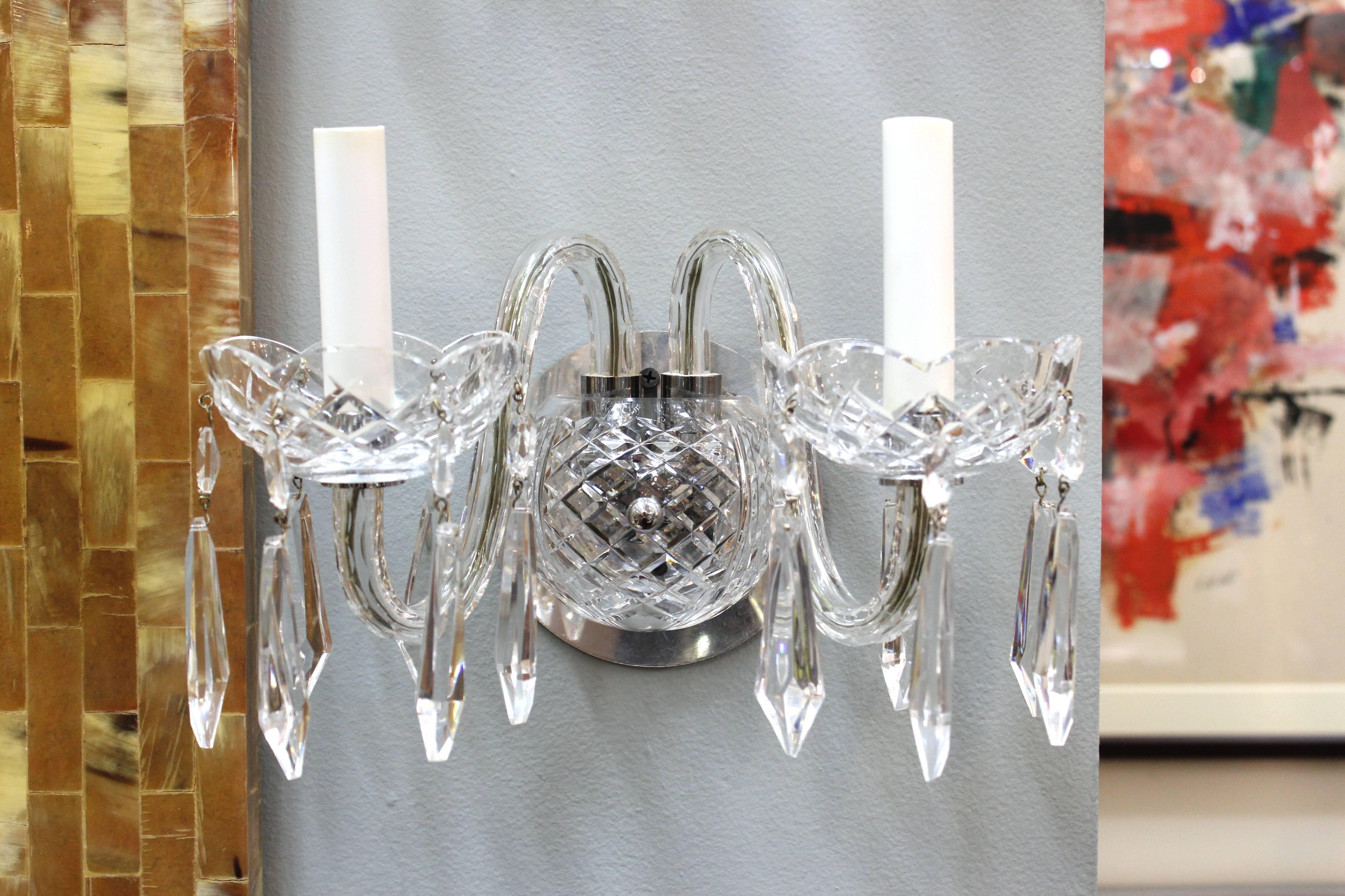 Set of sconces produced in Waterford crystal. Each piece features two carved arms attached to a cut crystal center. The scalloped faux candleholders are hung with faceted crystal drops. Minor wear consistent with use. In good condition.