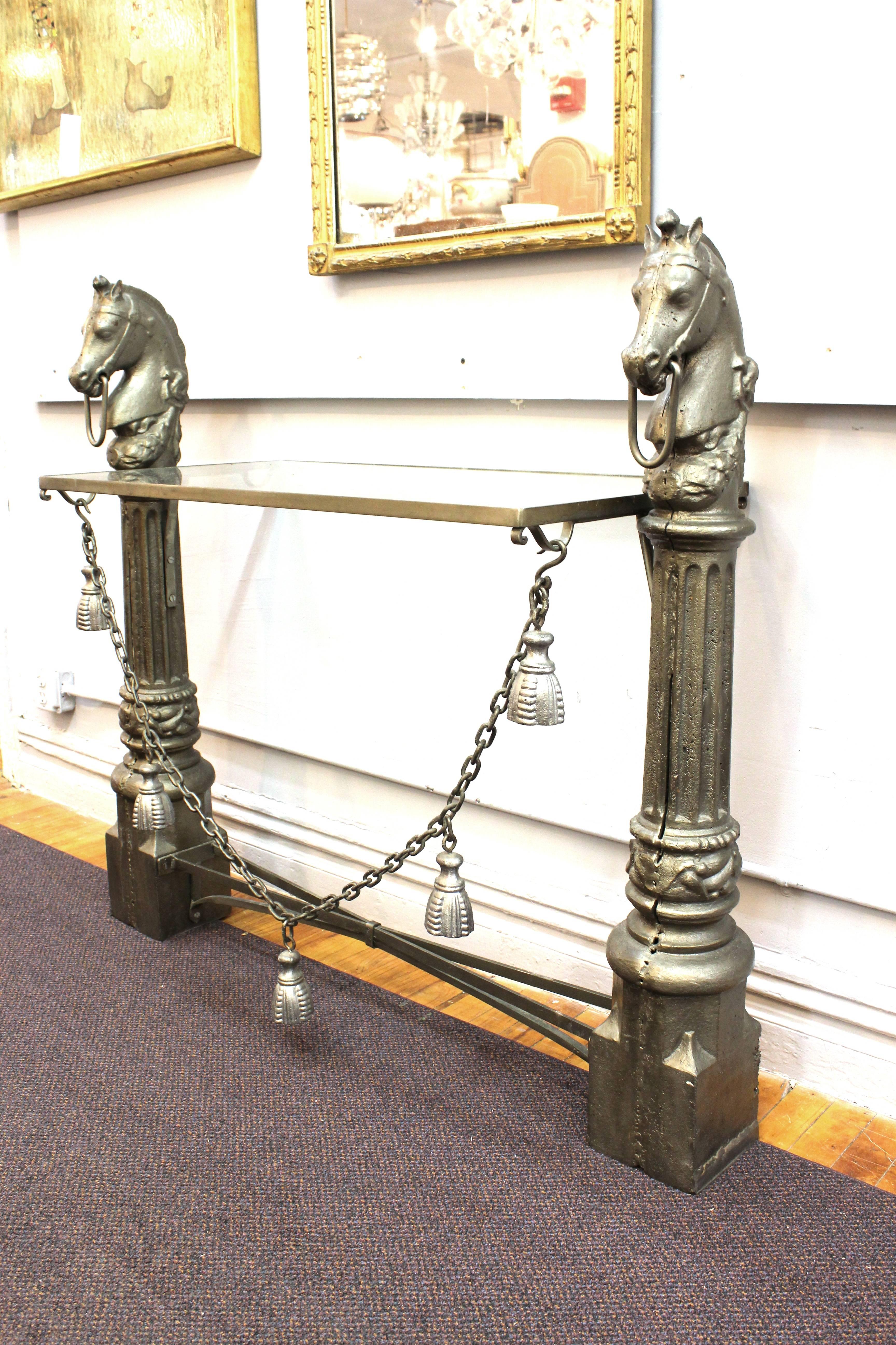 A metal sideboard designed to look like a pair of hitching posts with a horse head and column motif. Includes a glass shelf and a crossed bottom. The table features a long decorative chain with faux metal tassels. Despite wear consistent with use