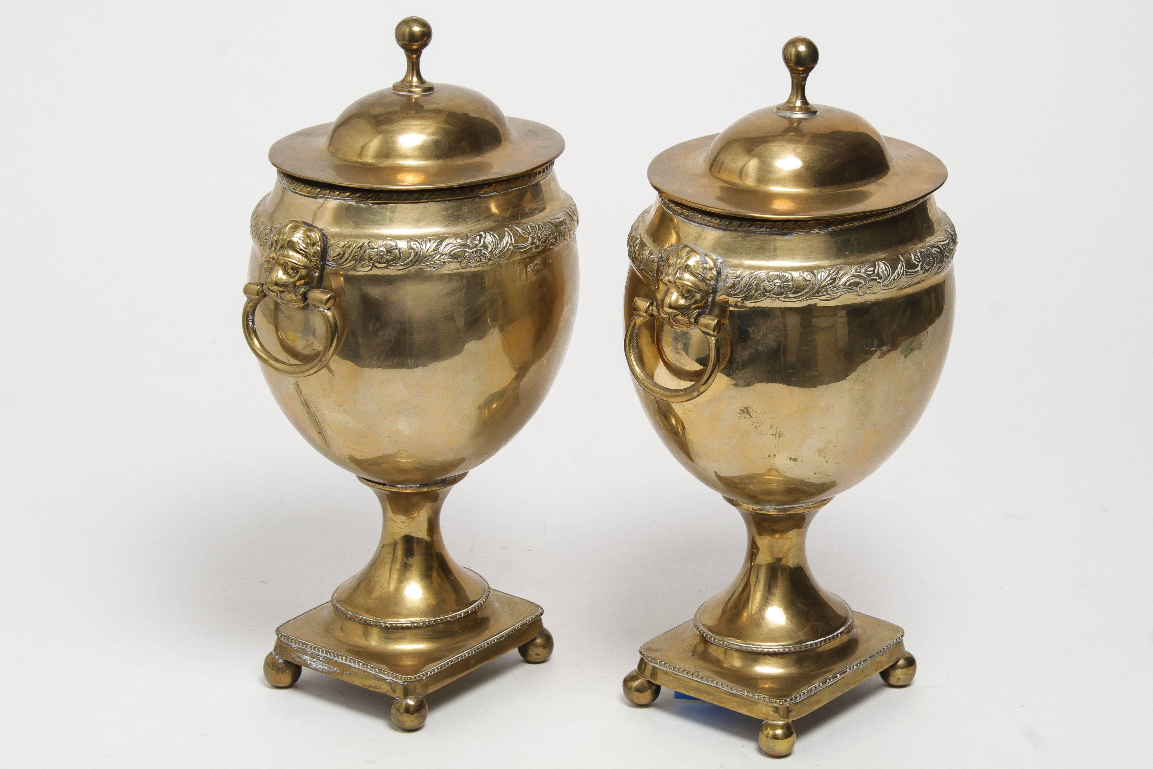A pair of footed solid brass urns with lids, made by Mottahedeh. A floral border runs along the top, with looped handles on each side, held by lion heads. Great vintage condition.