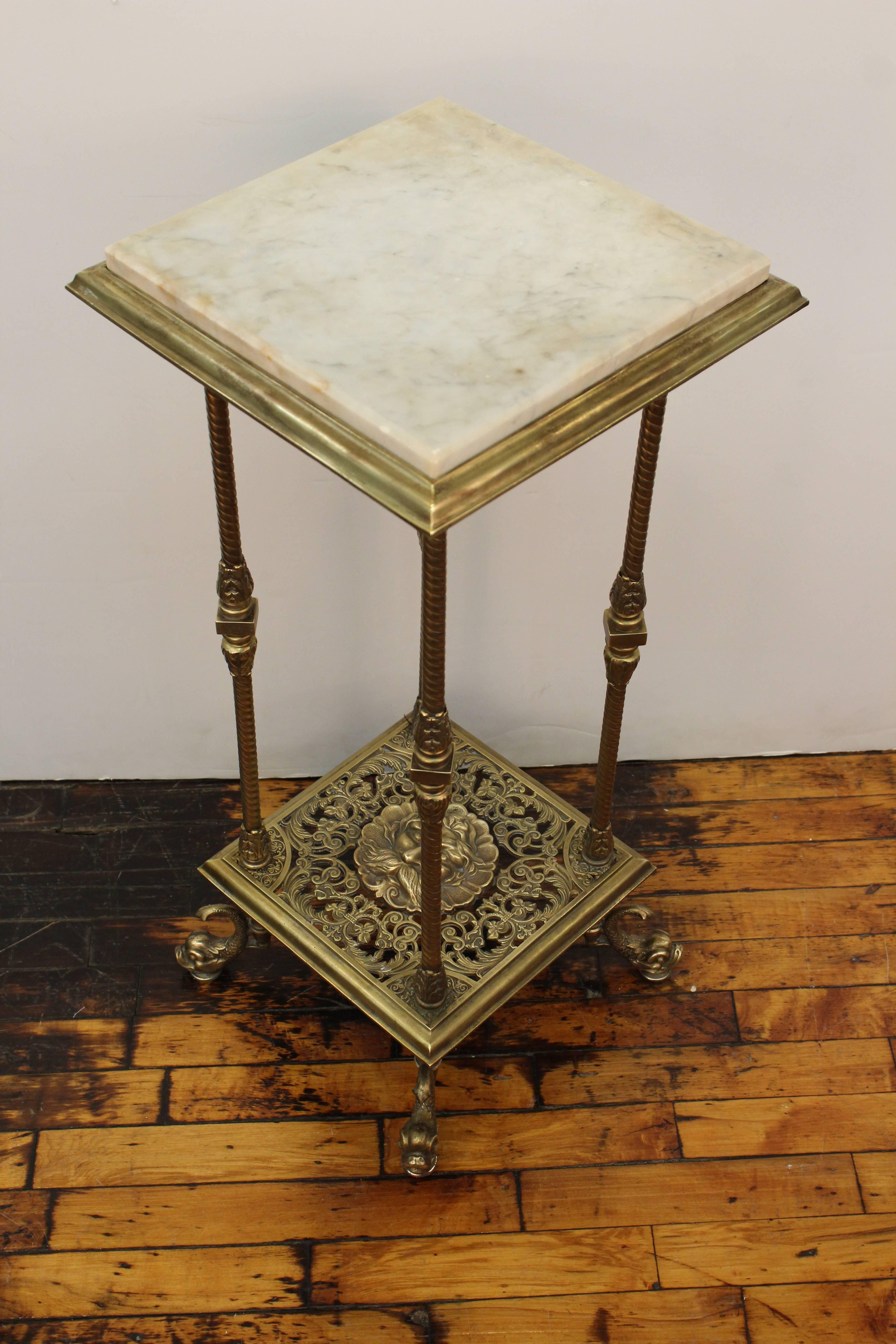 Victorian brass pedestal with elaborate decor and square marble top, made in the United States in the 1890s. The pedestal has sculpted dolphin feet and a decorative bearded mask on its lower tier. In very good vintage condition.