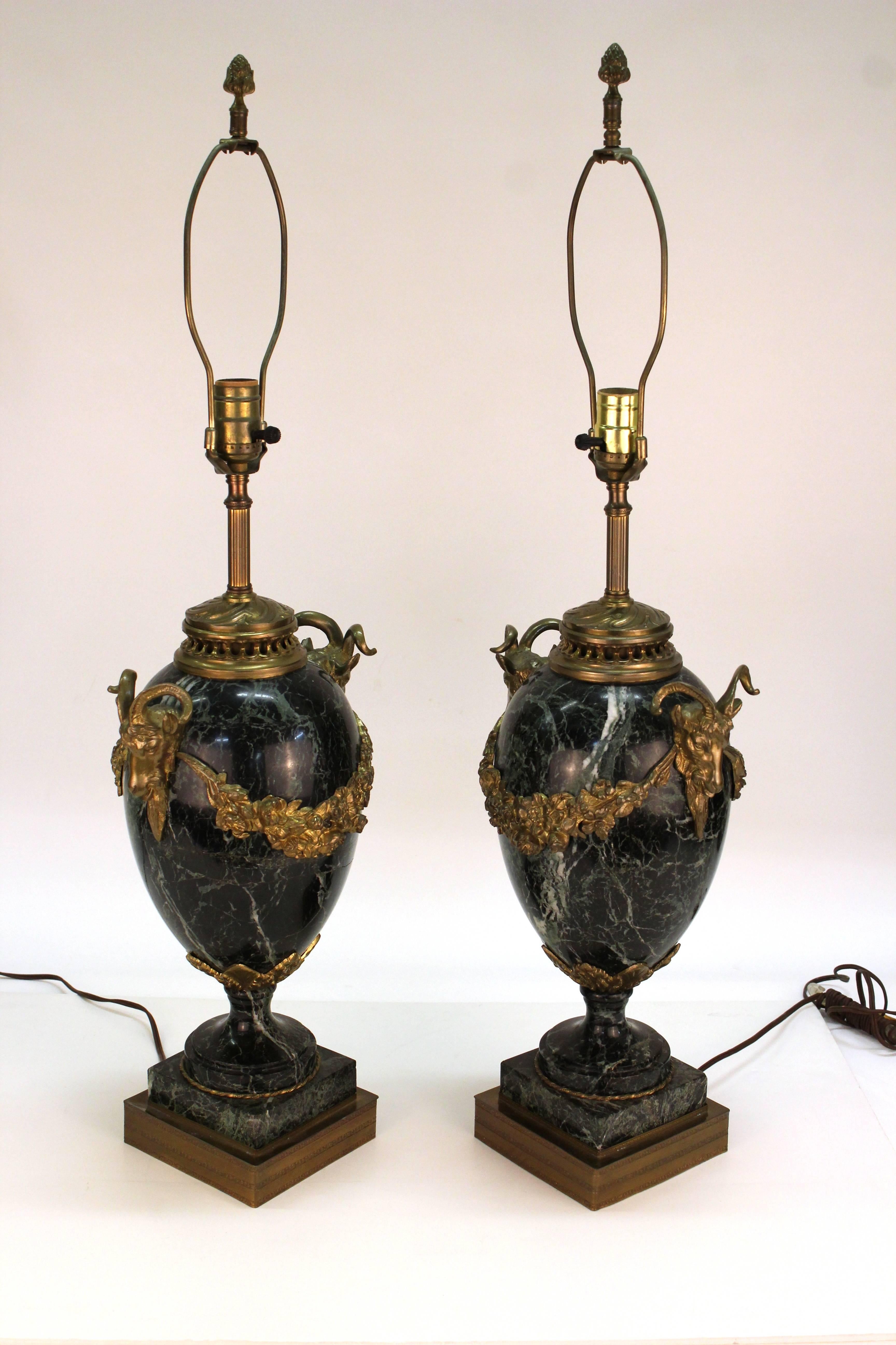 A pair of neoclassical marble table lamps in urn shapes. Sculpted marble with decorative accents of gilt bronze swags and ram's head handles on the sides. Slight chip to the marble on a corner of one of the bases. In overall good vintage condition.