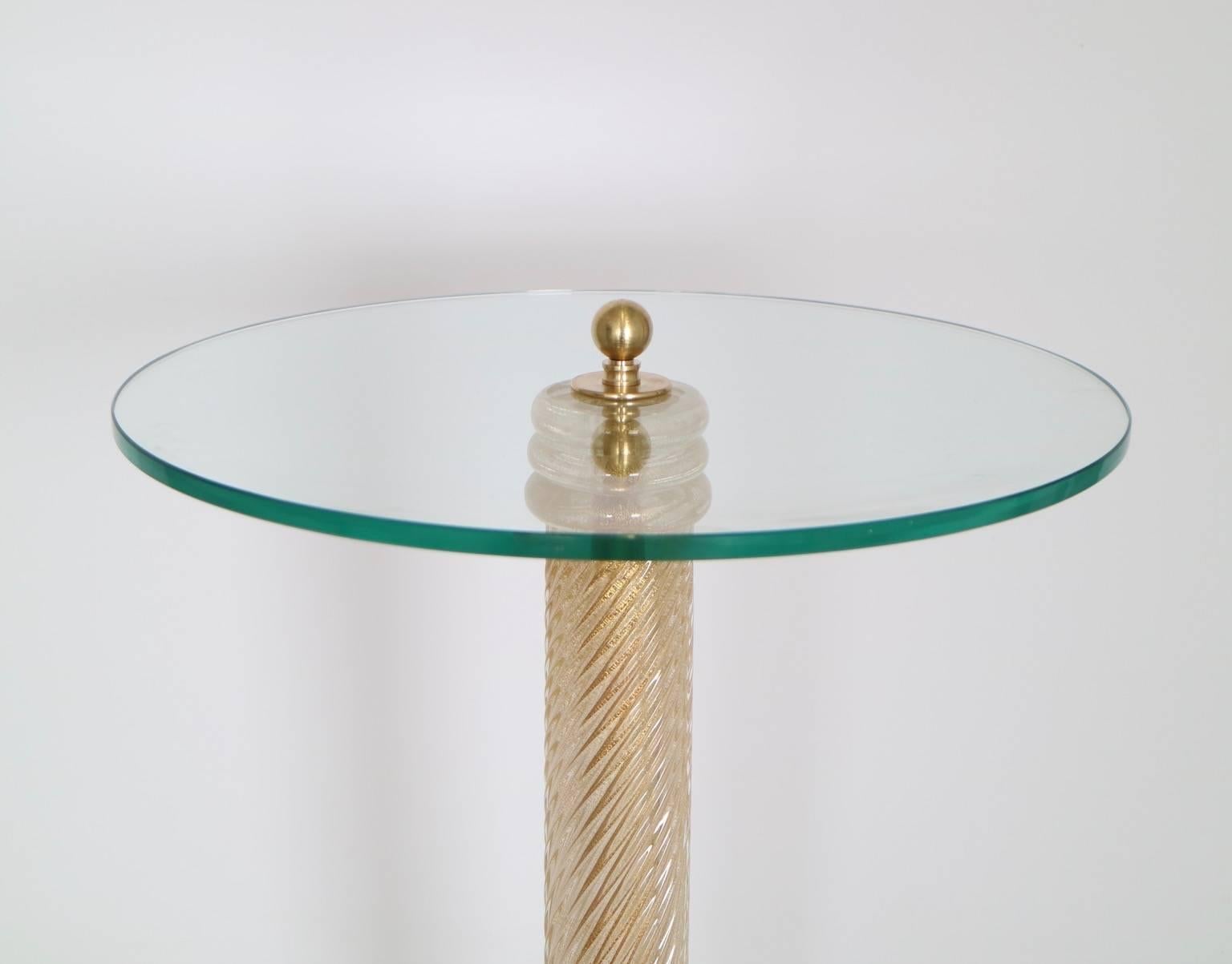 A Hollywood Regency Murano glass side table with circular top on Murano  glass pedestal with swirled gold flakes. The table is mounted on a circular gilded wooden base. The table top is in clear glass. This table is in excellent vintage condition