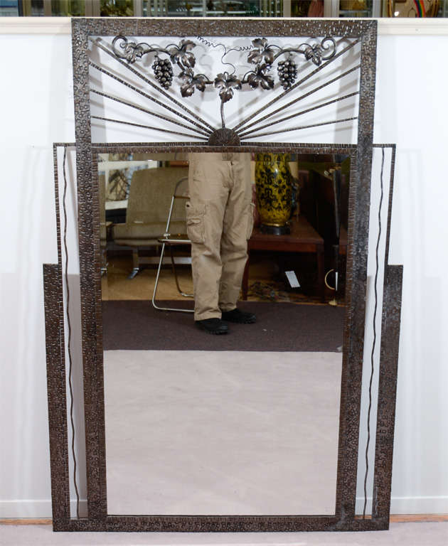 An original Art Deco mirror surrounded by a worked iron frame. The frame above the mirror is decorated with a grape vine with grapes and leaves. Good condition with age appropriate patina and wear; there is some spotting on the upper left side of