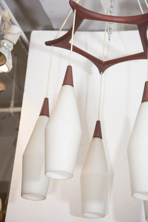 A vintage chandelier, produced in Denmark circa 1950s and 1960s by Holmegaard, with five milk glass cylindrical shades, each with a pointed teakwood top, suspended by white cords from a star-shaped teakwood stretcher. Markings include original