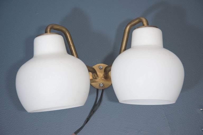 A pair of vintage Danish wall sconces, produced circa 1950s by designer Vilhelm Lauritzen and manufactured by Louis Poulsen, with double lights and bell-form milk glass shades, supported by curved brass arms and circular backplate. Good vintage