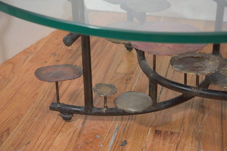 A vintage Brutalist style coffee table, designed by New York City based sculptor Silas Seandel, circa 1970s, with a round glass top, above a varied sequence of stylized, mixed metal lily pads, each disk affixed to alternating support beams as base.