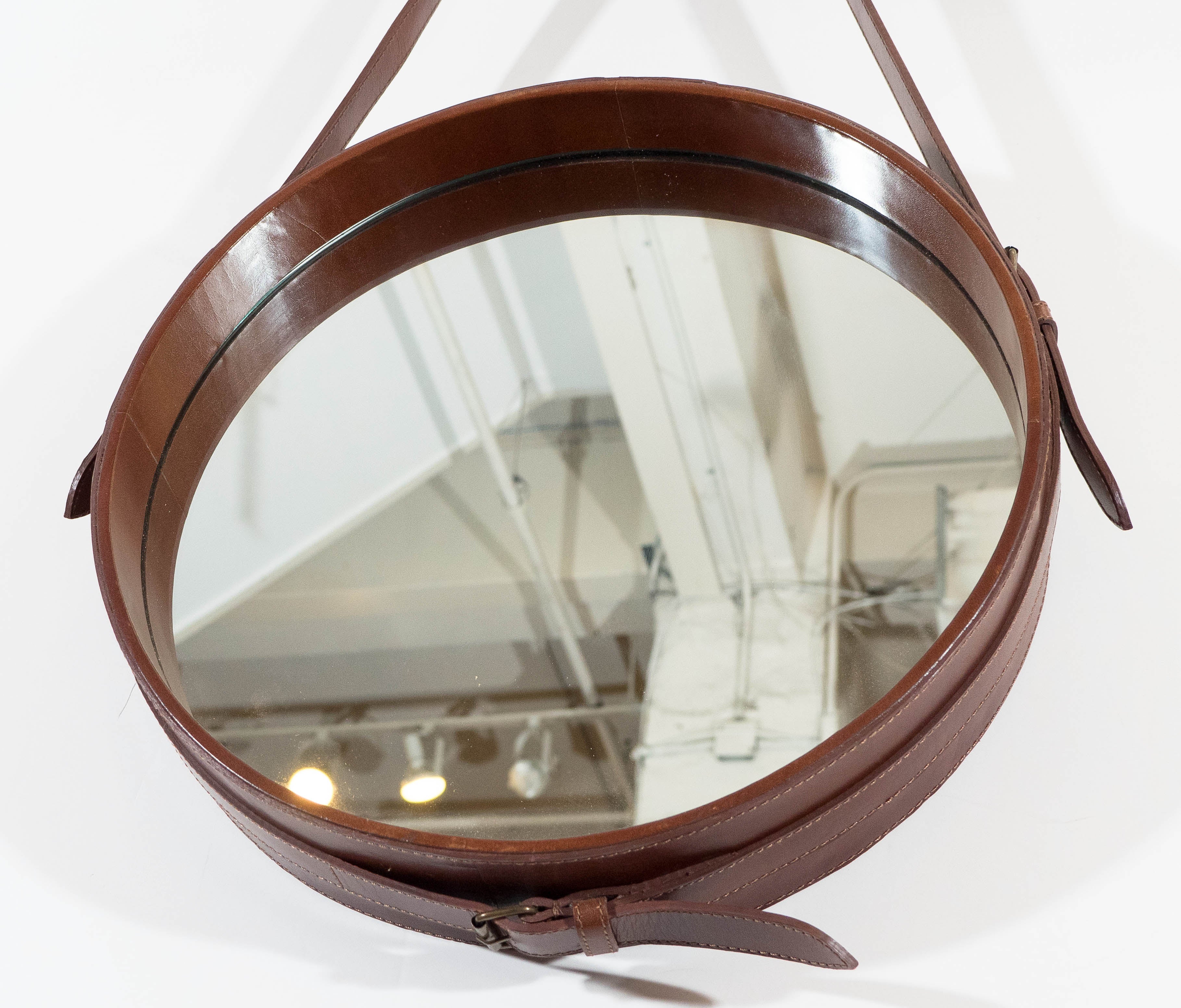A round Adnet wall mirror, based on the original designs by namesake Jacques Adnet, the frame and strap-work in faux brown leather, with brass buckles. Very good condition, wear consistent with age and use; nice patina to the brass.