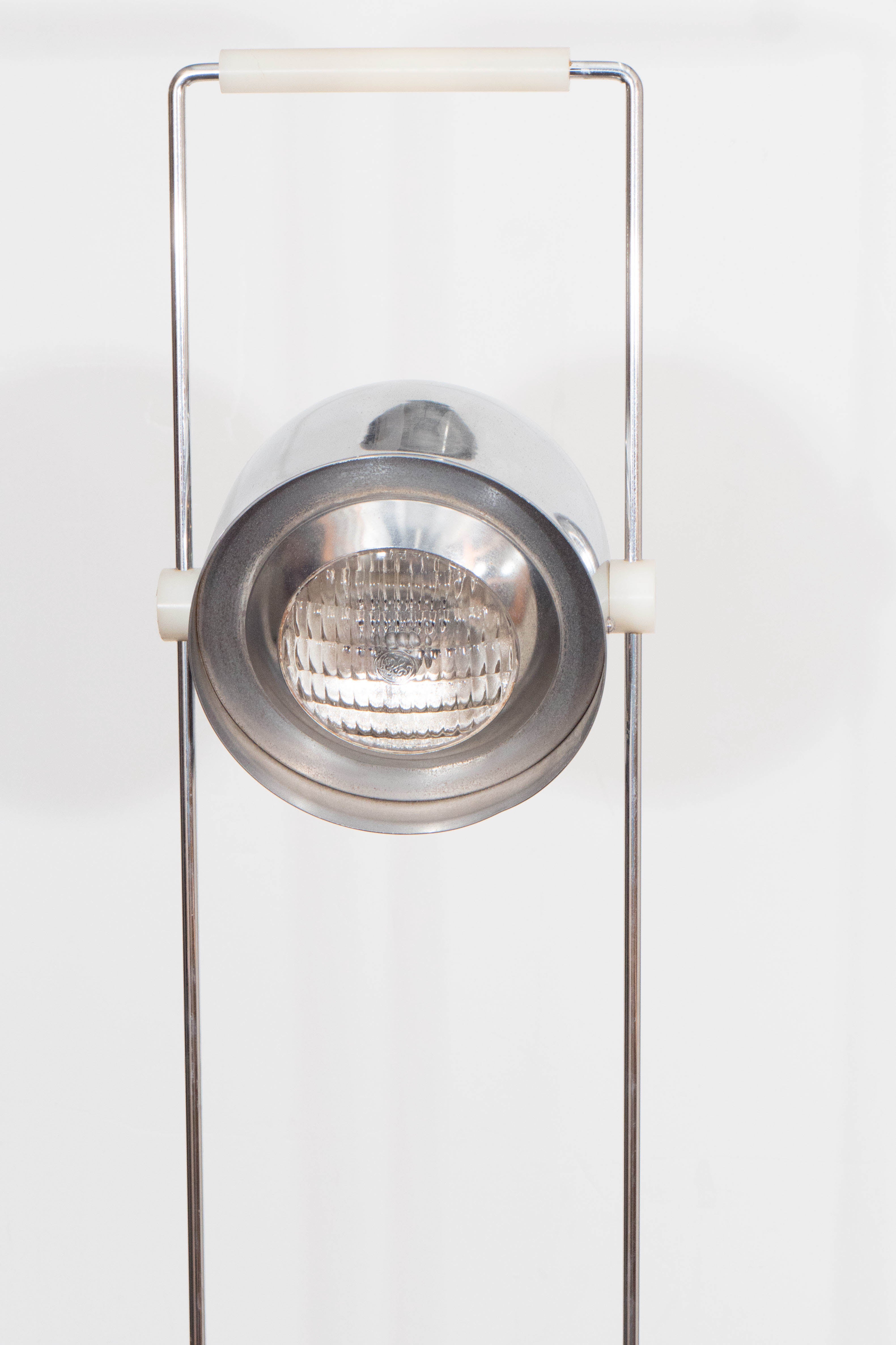 An adjustable spotlight as floor lamp, produced in the United States circa 1970s, with two lights in chrome shades, supported by a long angular frame on a cylinder base; includes on/off switch on cord. Wiring to US standard. Good condition, with