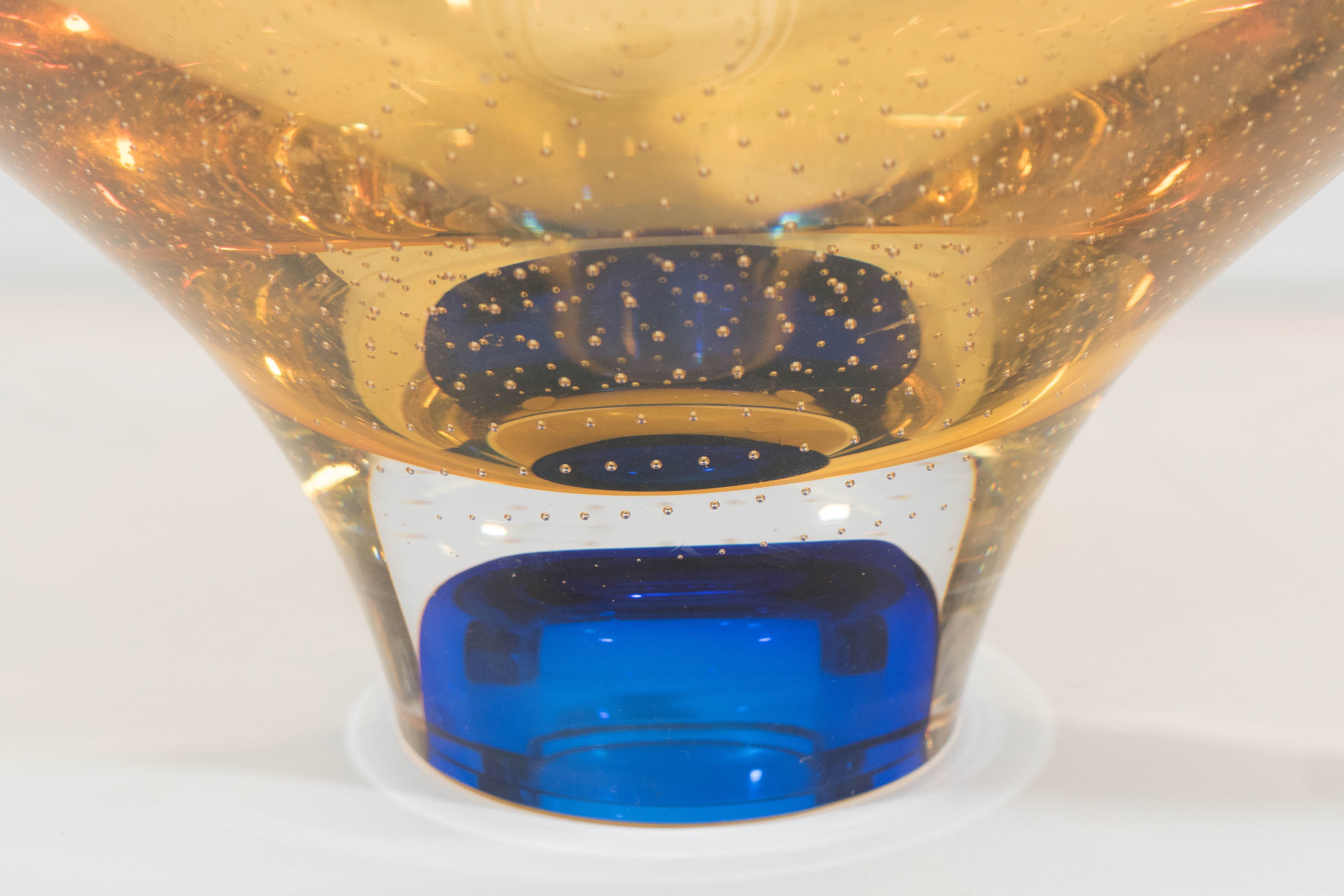 A decorative amber colored art glass bowl, with conical oval shape on cobalt base, with the inclusion of small controlled bubbles, by Swedish designer Göran Wärff, produced by Kosta Boda, Sweden. Markings include: inscription on the bottom ‘Kosta