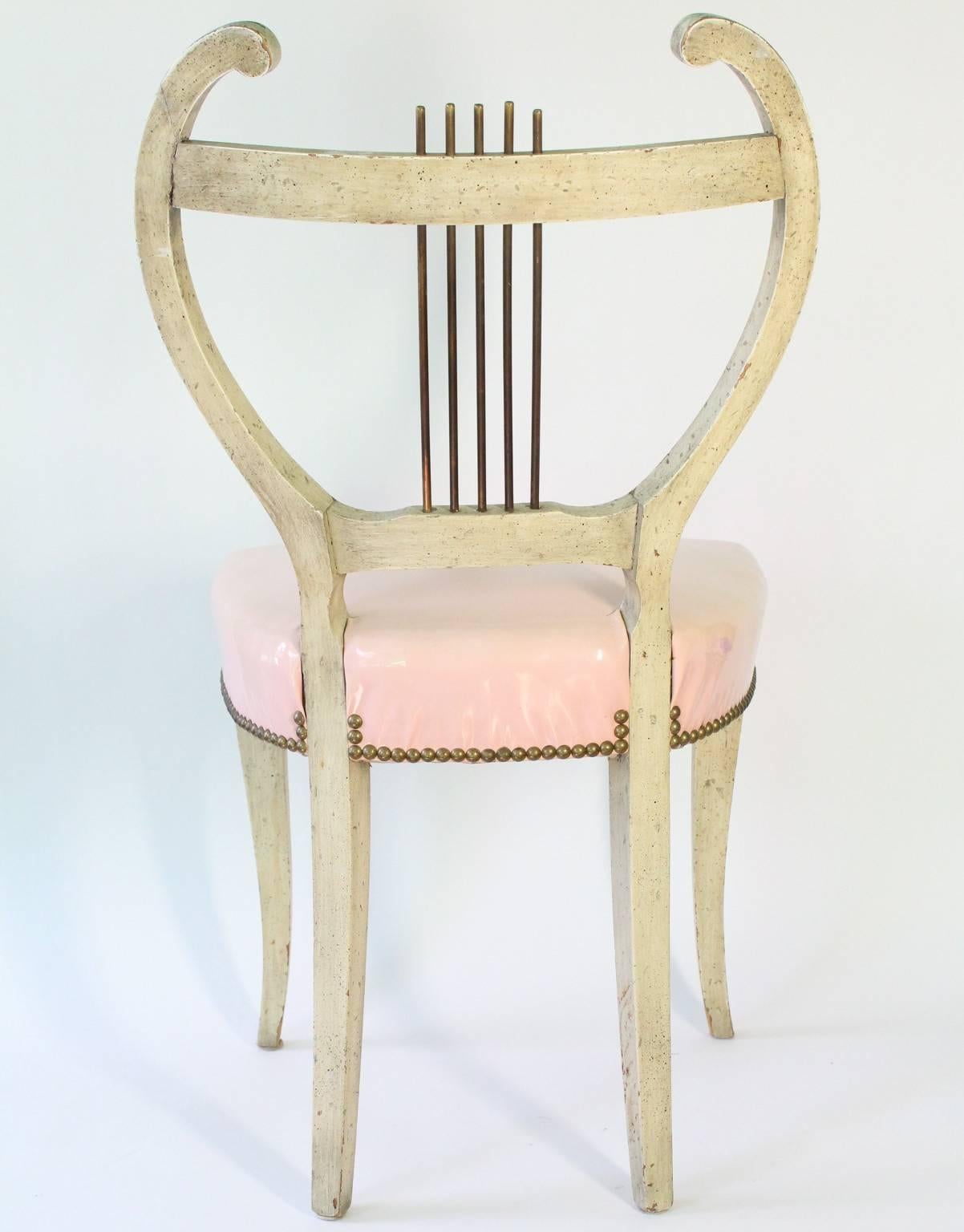 A set of four French vintage dining chairs, designed in the Regency style and produced circa 1950-1960, each with painted wood frames on Klismos legs and pink vinyl seats, lined with brass nailhead studs, the stylistic lyre backs accented with brass