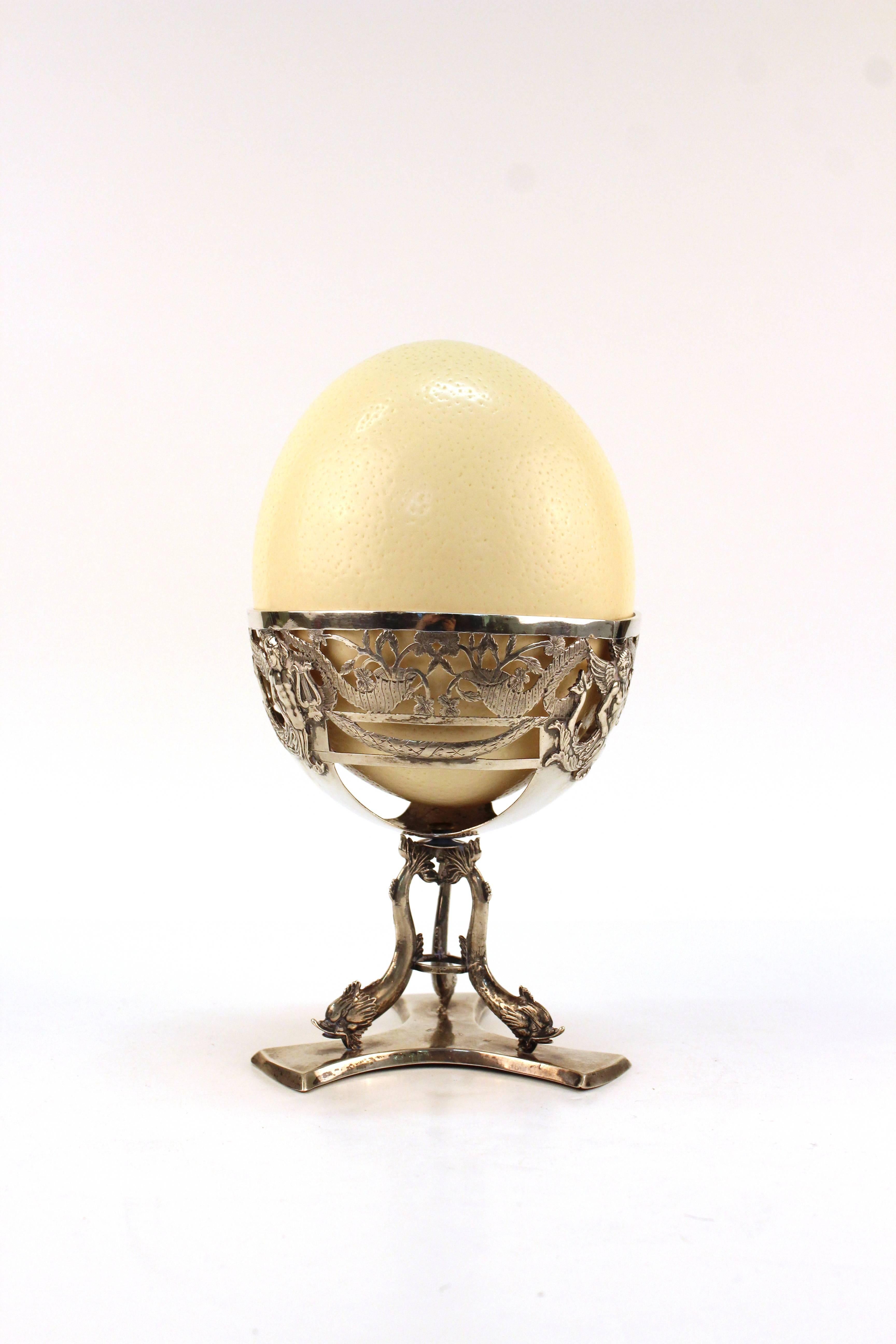 An antique ostrich egg placed in a German silver stand. The tripod base decorated with dolphins while the Stand features mythological beings and cornucopias. Stamped on the base. Despite some tarnish and wear the piece remains in good vintage