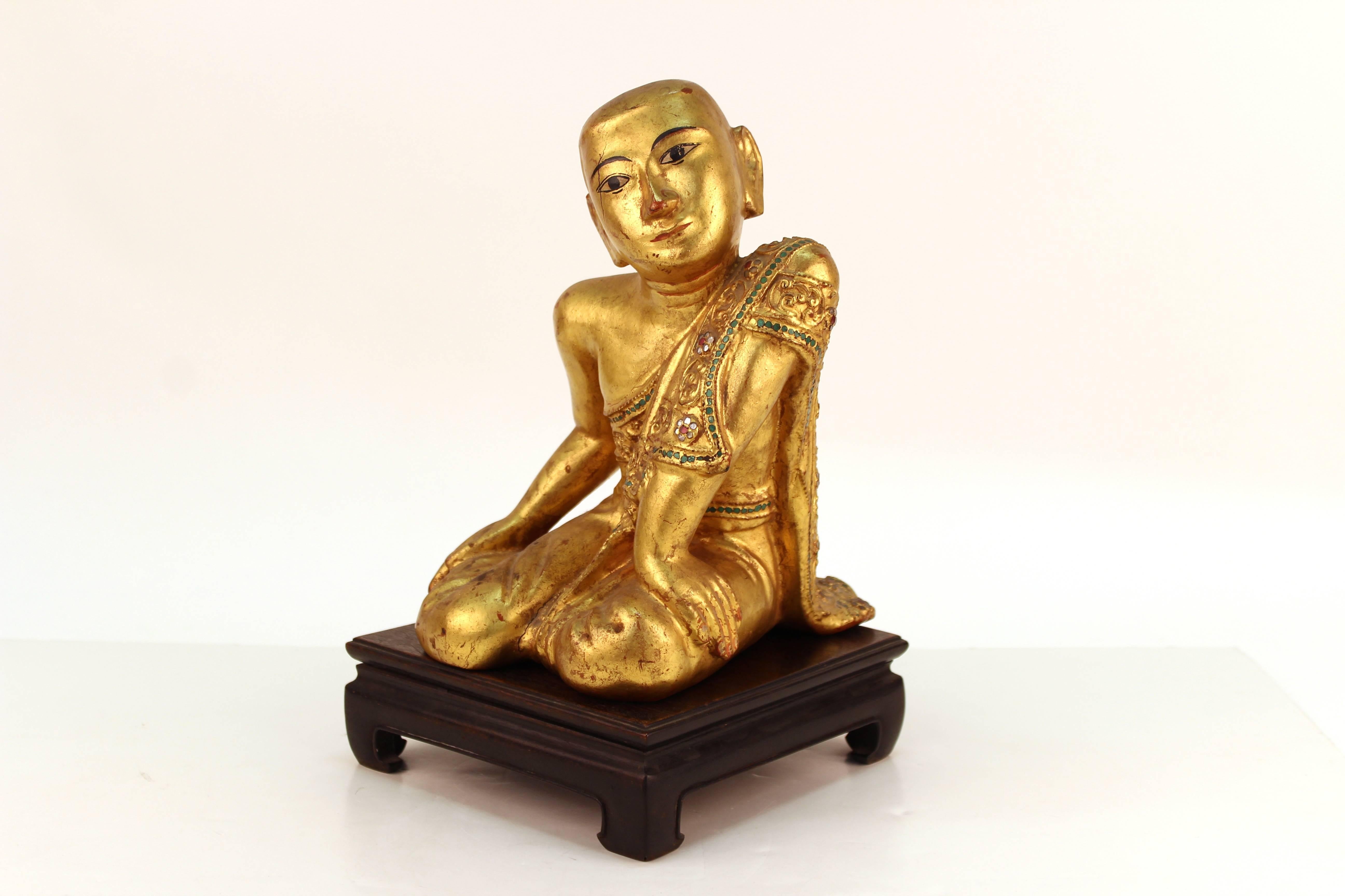 Buddhist monk sculpture from Myanmar (previously Burma). Crafted from carved giltwood. Depicts a young monk in traditional dress. The figure is seated comfortably on his knees leaning on one arm as he looks thoughtfully to the side. The sculpture is