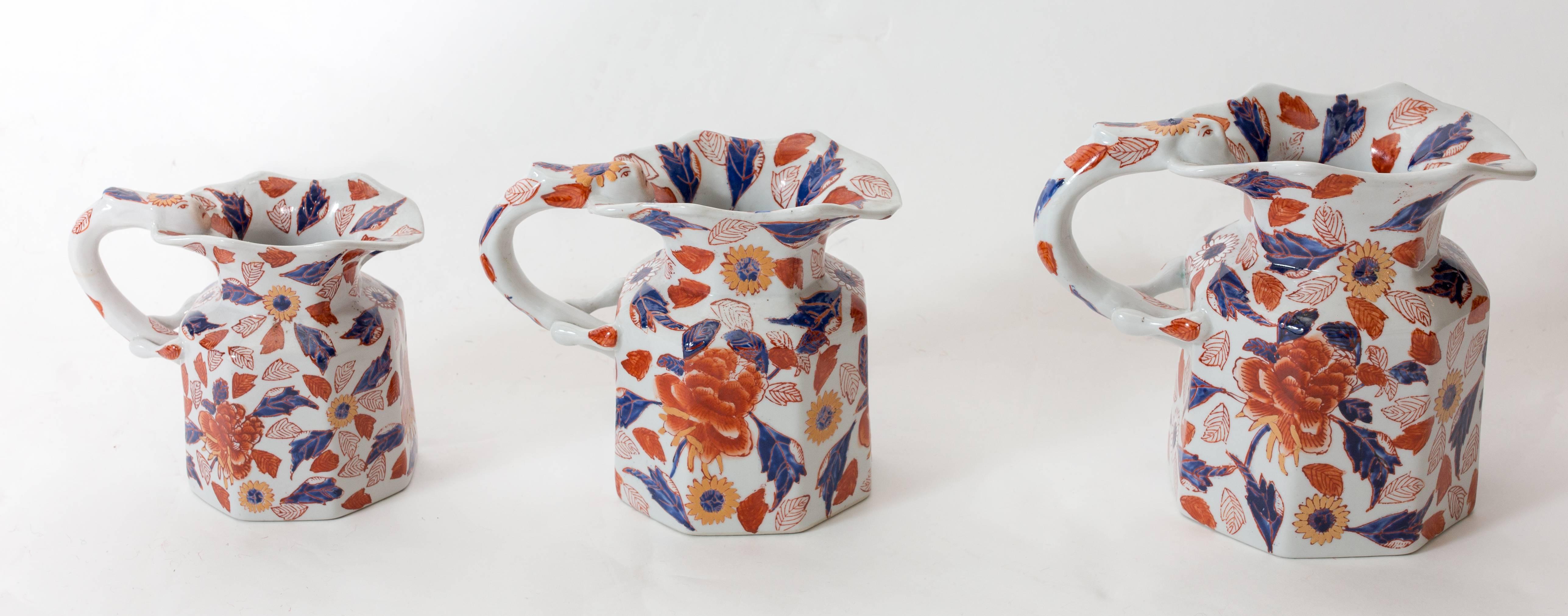 Beautiful trio of pulled handle, handmade, hand thrown porcelain pitchers each with a face motif atop the handle with hand-painted floral glaze of purple, orange and gold.