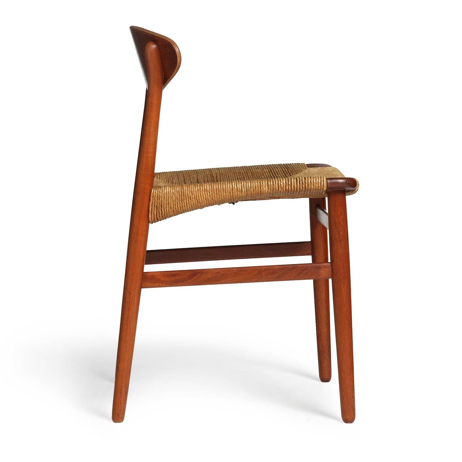 Una poltroncina in teak sobria ed elegante, con schienale curvo e fluttuante, seduta in giunco intrecciato e un espressivo dettaglio a tenone passante sulle gambe anteriori.
