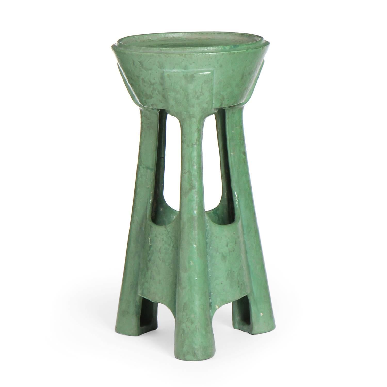 An interesting architectural ceramic stand or end table having a tapered top supported by three flaring organically shaped legs, the entire form covered in a rich and complex matte green glaze.