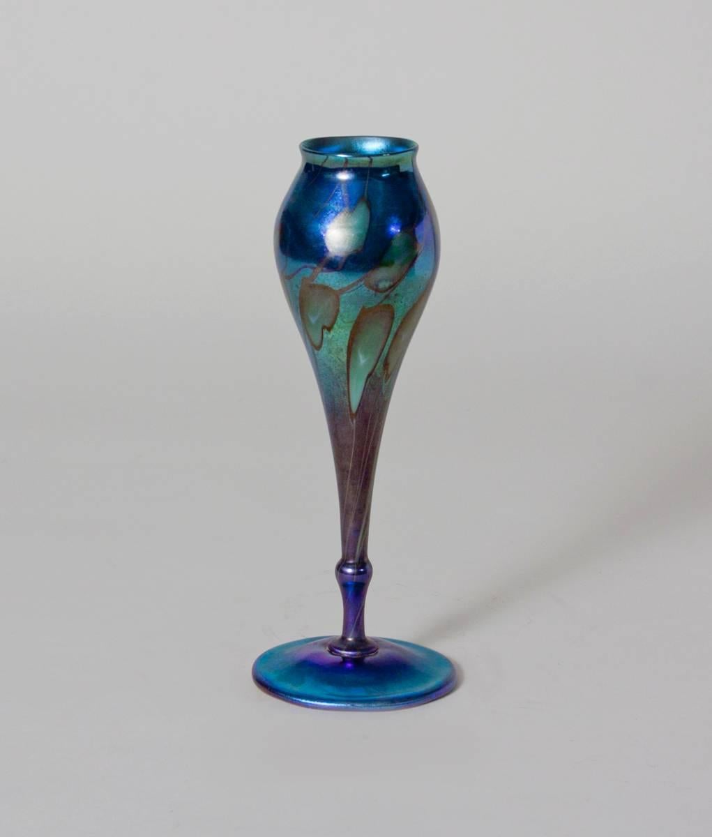 A Tiffany Studios Favrile Glass Flower Form Vase comprising an elongated form in a deep iridescent blue decorated with leaves and vines, signed.