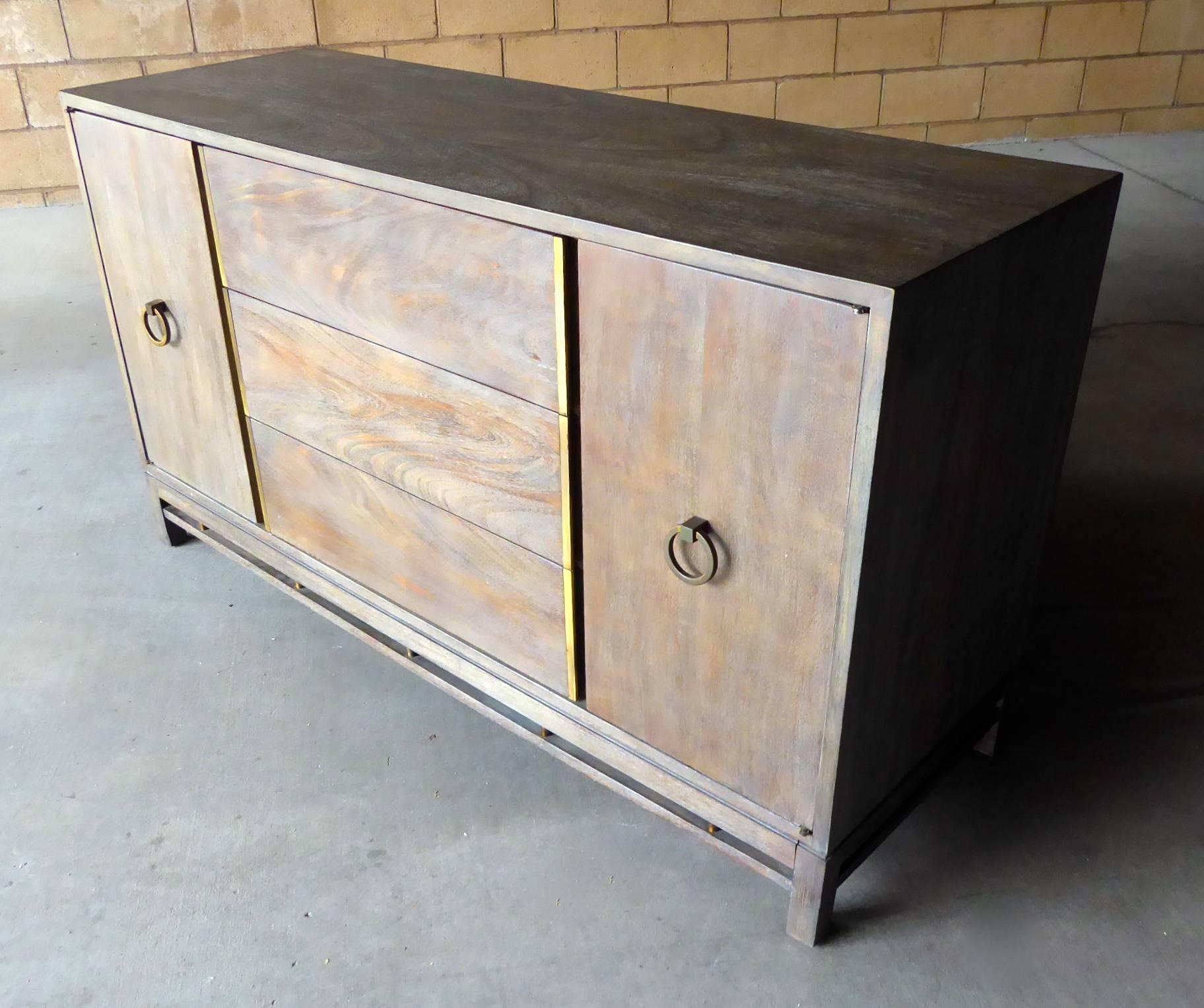 A chic stained walnut 1960s credenza by American of Martinsville. The finish on the cabinet has been customized by the addition of a grey dry-brush technique over a medium toned walnut stain. The brass hardware is original and retains it toned-down