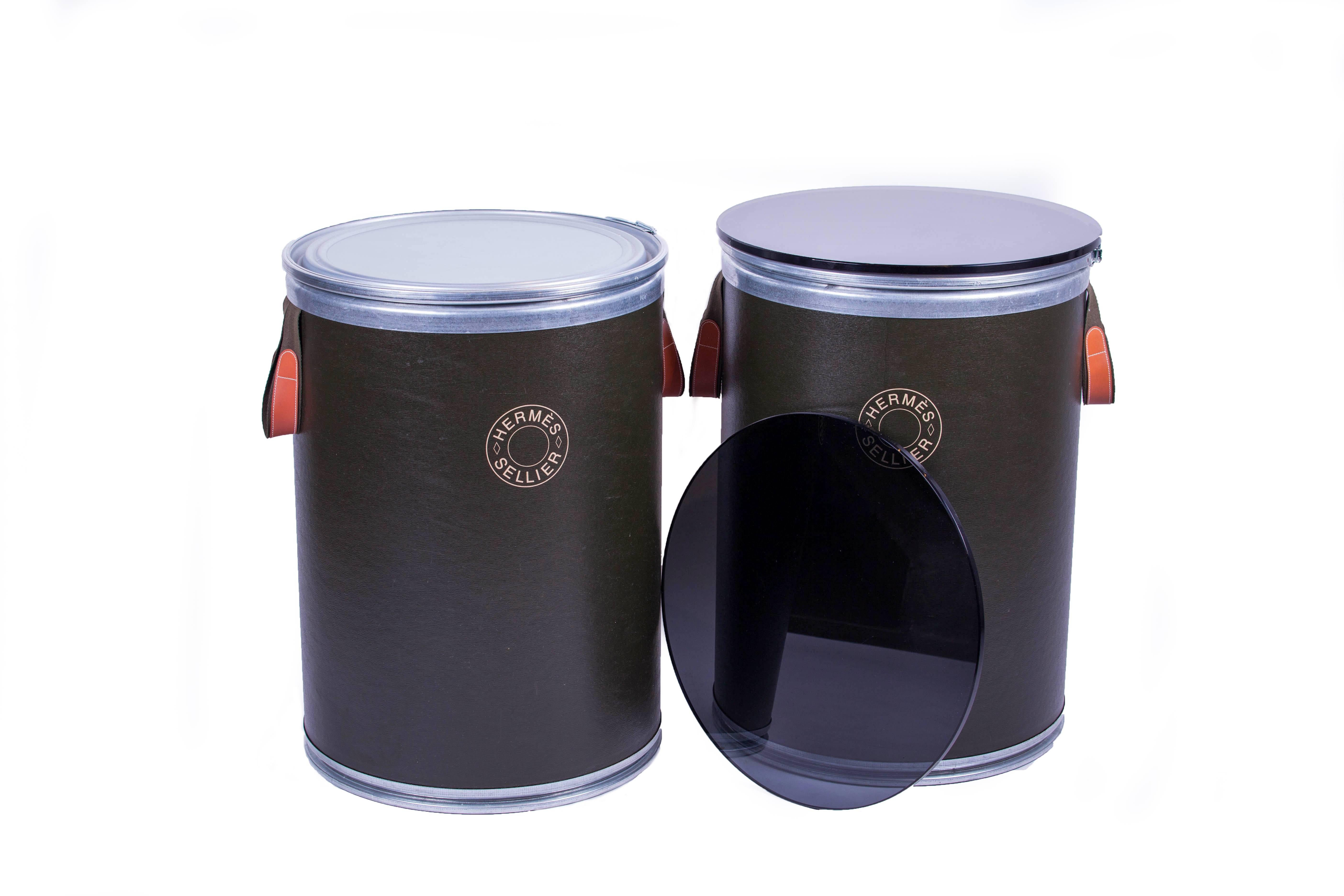 A creative pair of glass topped metal and board circular side tables from Hermes. Initially used as containers to transport the high-end saddles made by Hermes, this pair has been repurposed as side tables with the addition of the smoked glass tops.