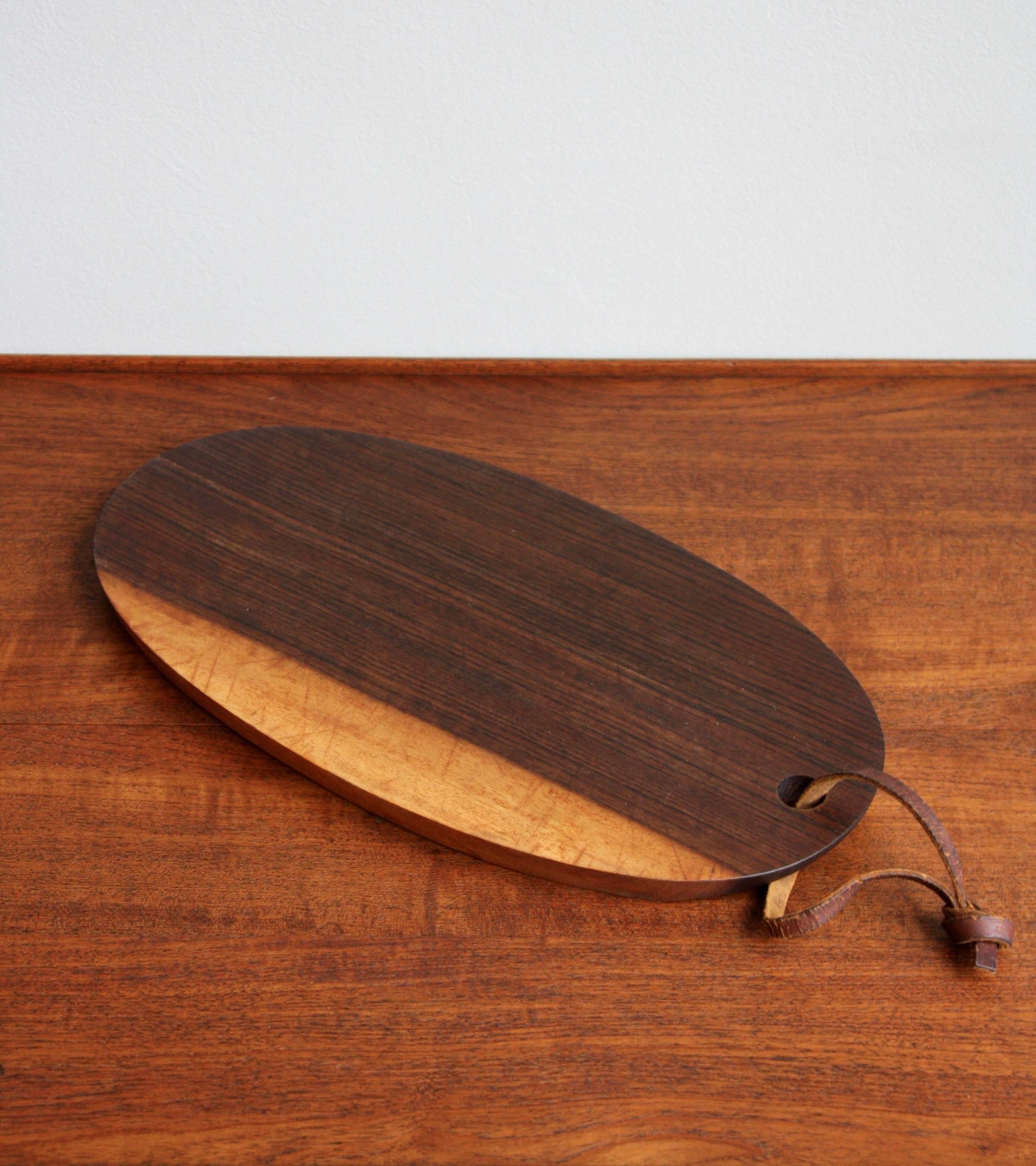 A vintage cutting board designed and made by Carl Auböck II. 
The elliptical shaped board is made from solid walnut. The ends of each side are geometrically chamfered to make it easier to grab and lift the board. 
At one end of the platter a