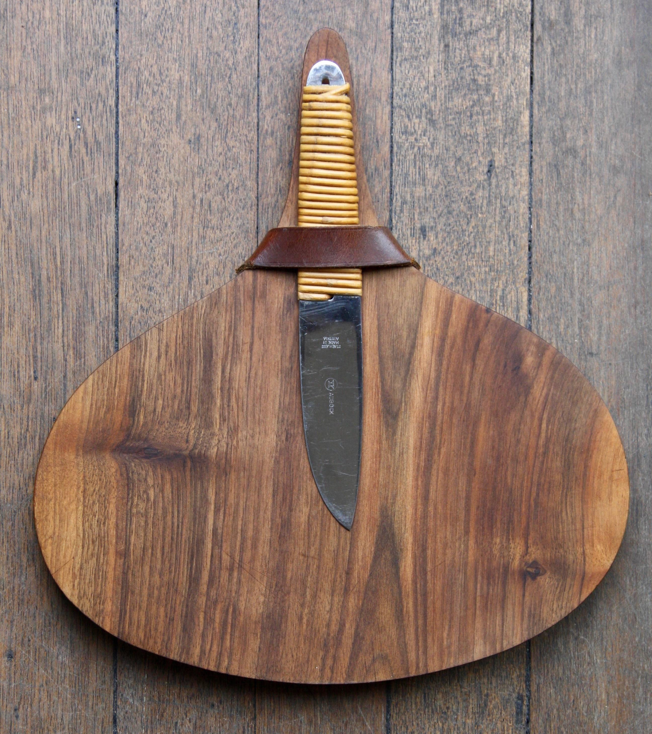 A vintage cutting and serving board with knife designed and made by Carl Auböck II, Vienna, circa 1950.
The board is sculptured from solid walnut and features a handle so that items may be presented on the platter. A dark tan brown leather strap